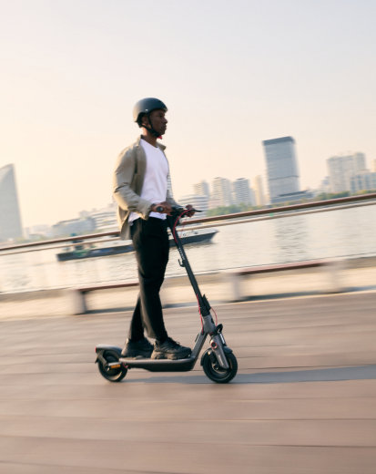 Homme conduisant une trottinette électrique sur une promenade. Bâtiments et eau derrière.