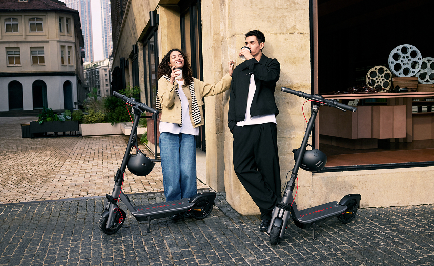 Un homme et une femme avec des scooters se tiennent dans une rue de la ville, tenant des tasses. Les scooters sont noirs et rouges.