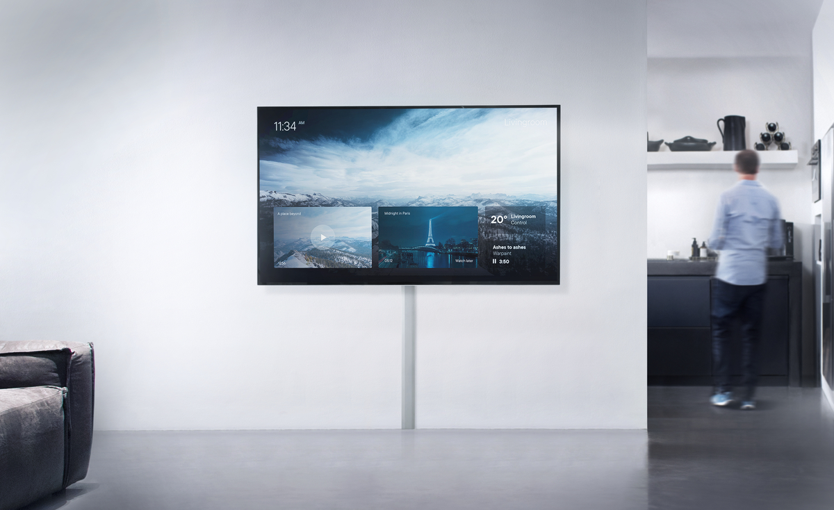 Ein großer Fernseher an einer weißen Wand, der ein Menü zeigt. Eine Person geht im Hintergrund.