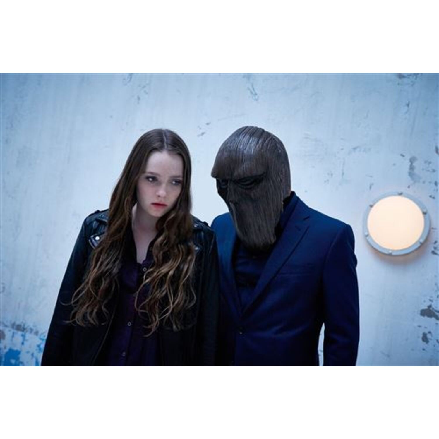 A man in a suit and a person in a mask stand together in front of a light blue wall.