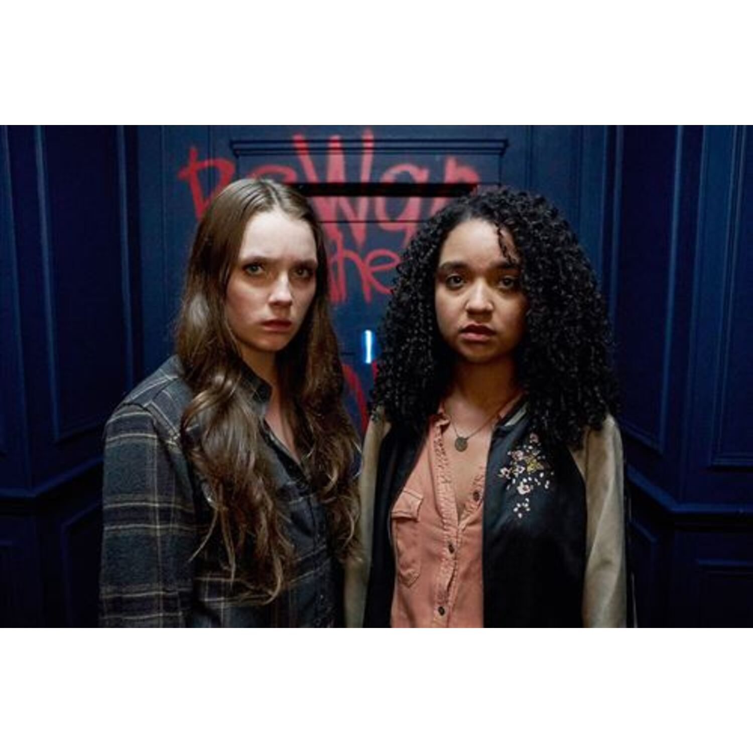 Two women stand in a dark room. Graffiti on the wall says "beware the". Both are looking at the camera.