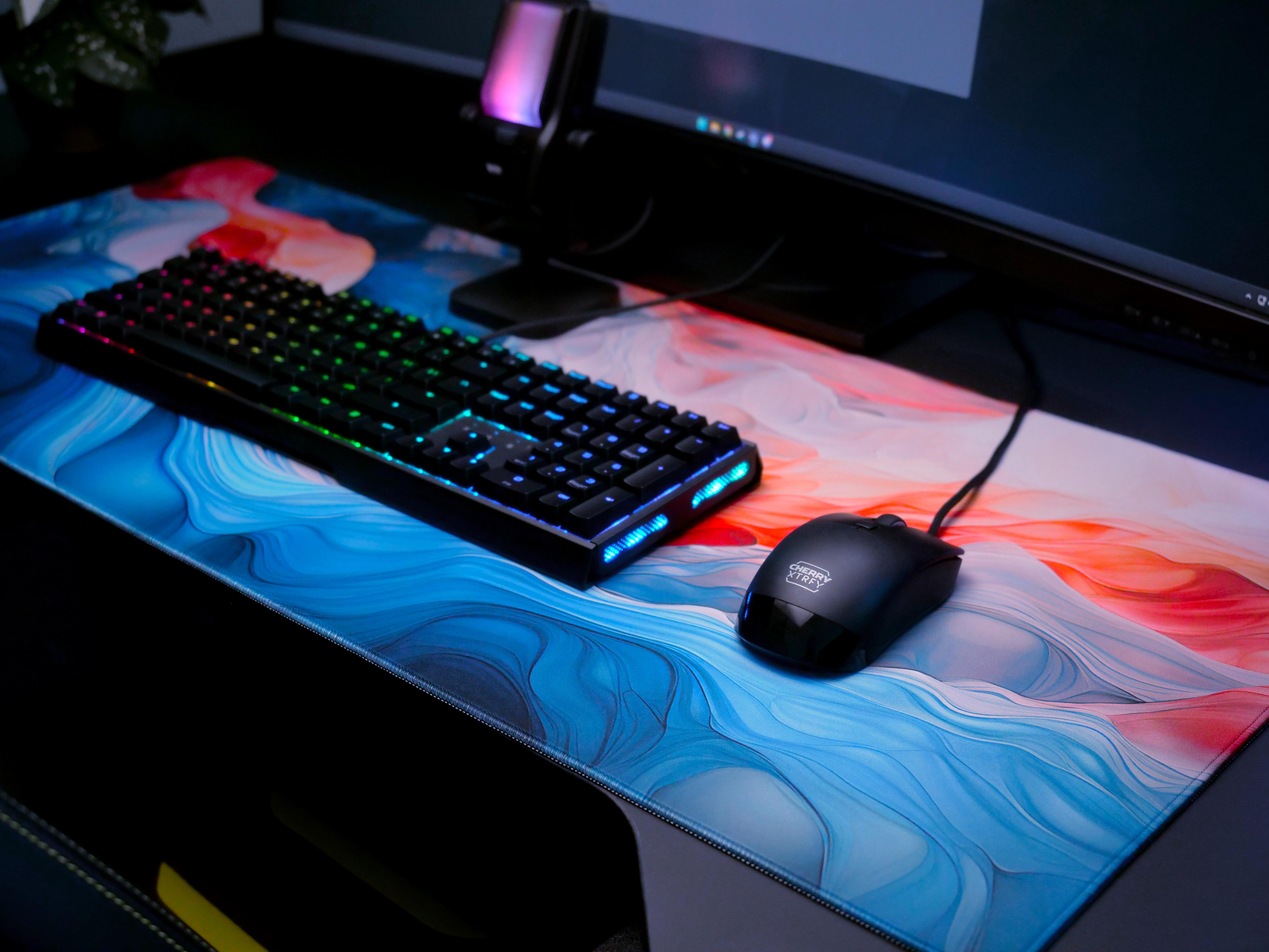 Gaming-Setup: Tastatur, Maus und bunte Matte. Hintergrund enthält Monitor und Schreibtisch.