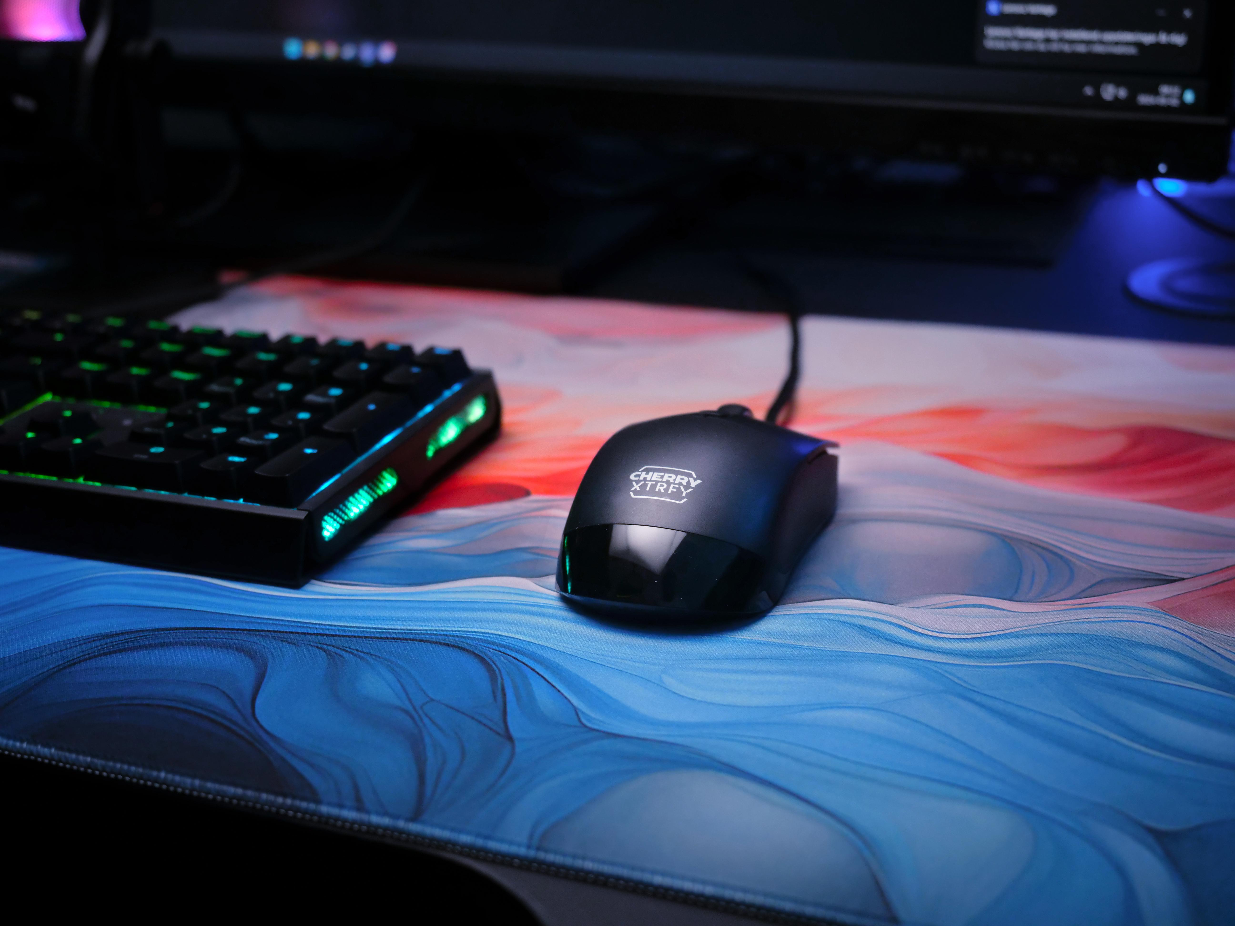 Ein Gaming-Setup. Eine schwarze Maus und Tastatur auf einem bunten Mousepad. Ein Monitor ist im Hintergrund.