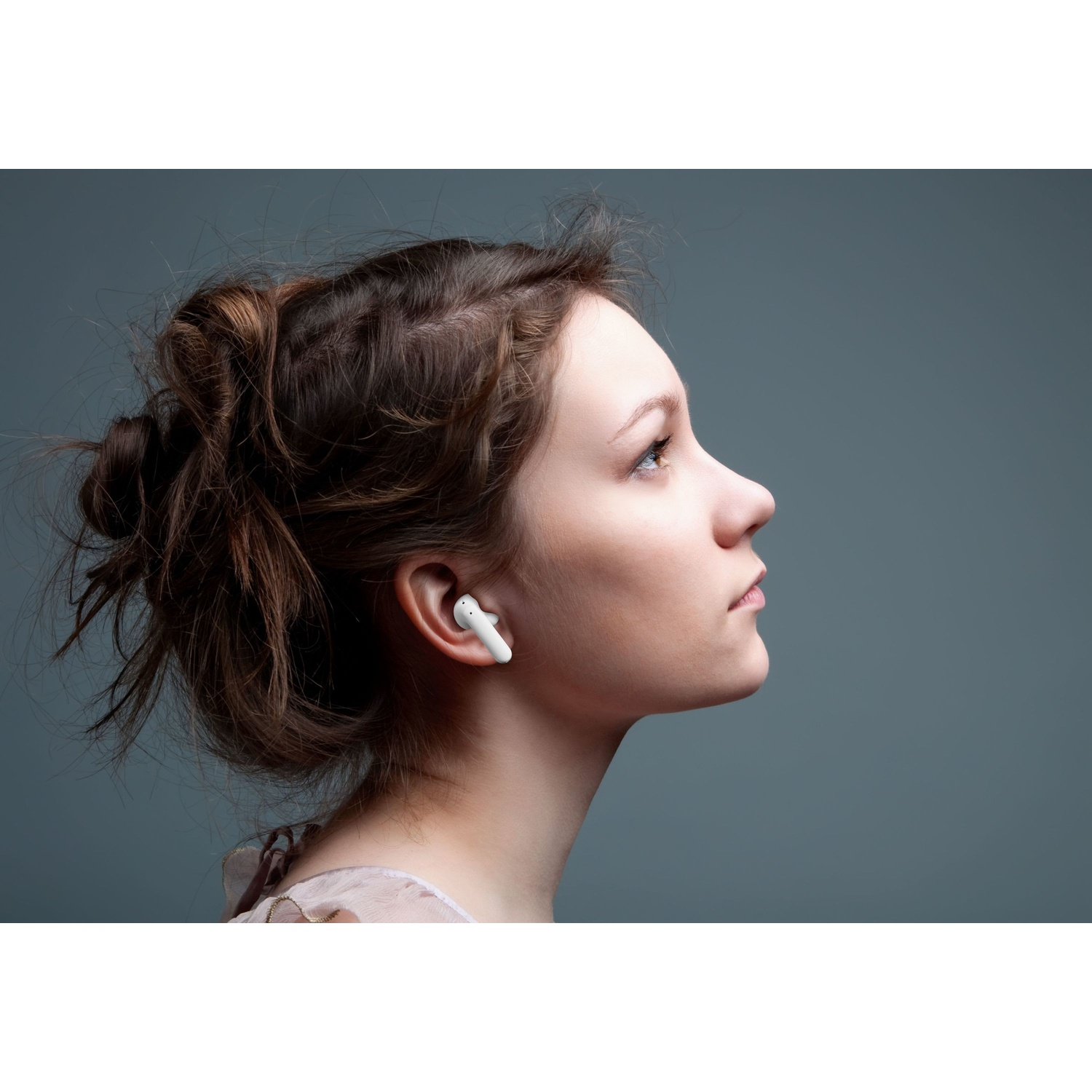 Una donna con i capelli castani e un auricolare bianco guarda in alto su uno sfondo grigio.