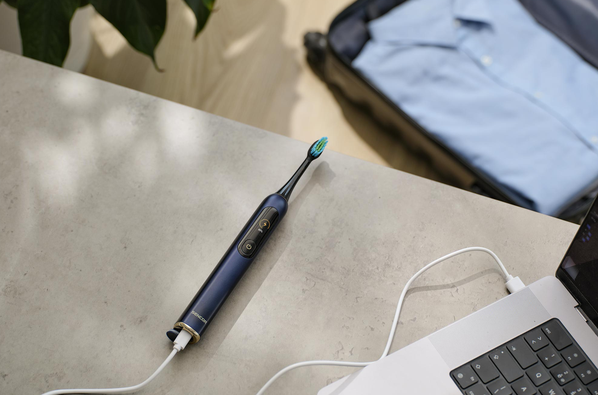 Kék fogkefe laptophoz csatlakoztatva, a háttérben egy bőrönddel.
