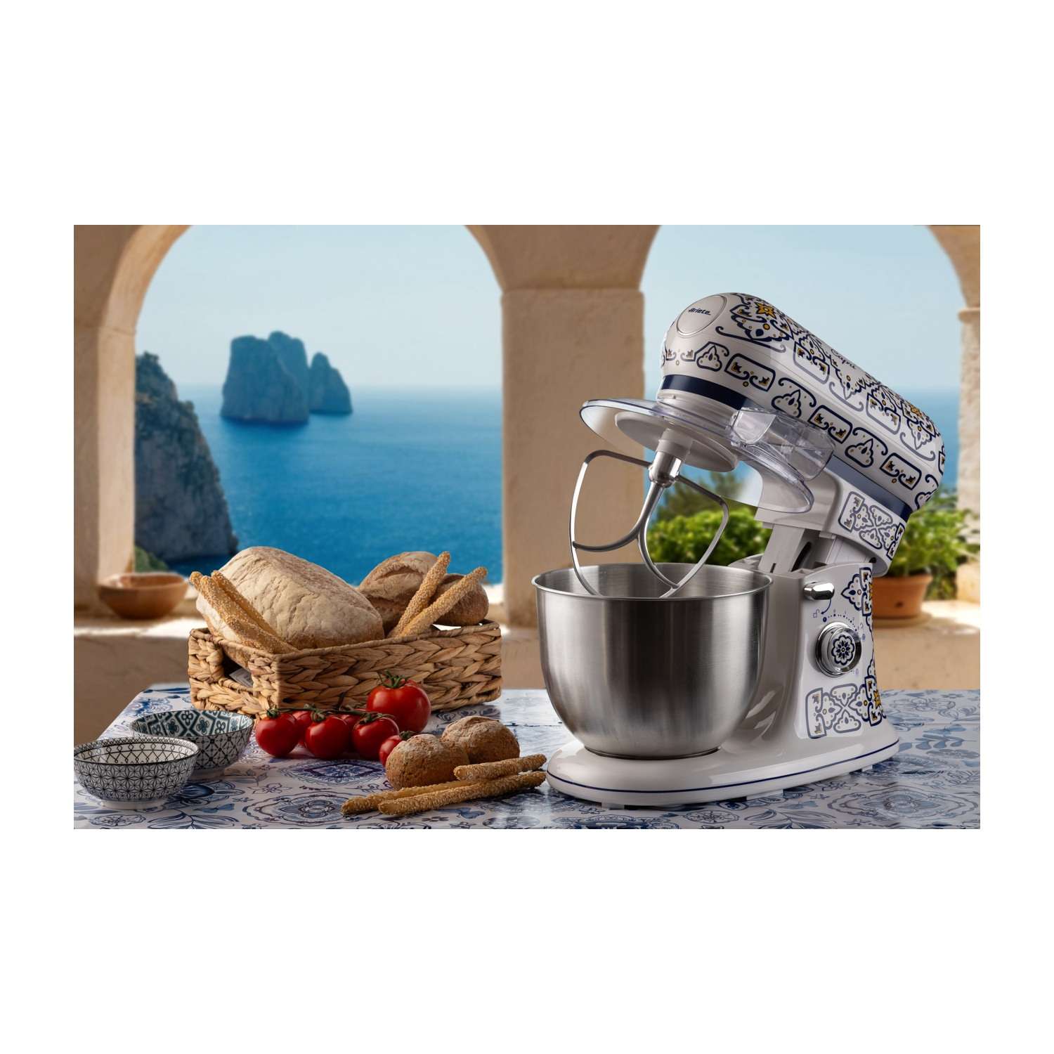 Un mixer da cucina con cibo e vista mare sullo sfondo. Un cesto di pane e pomodori.