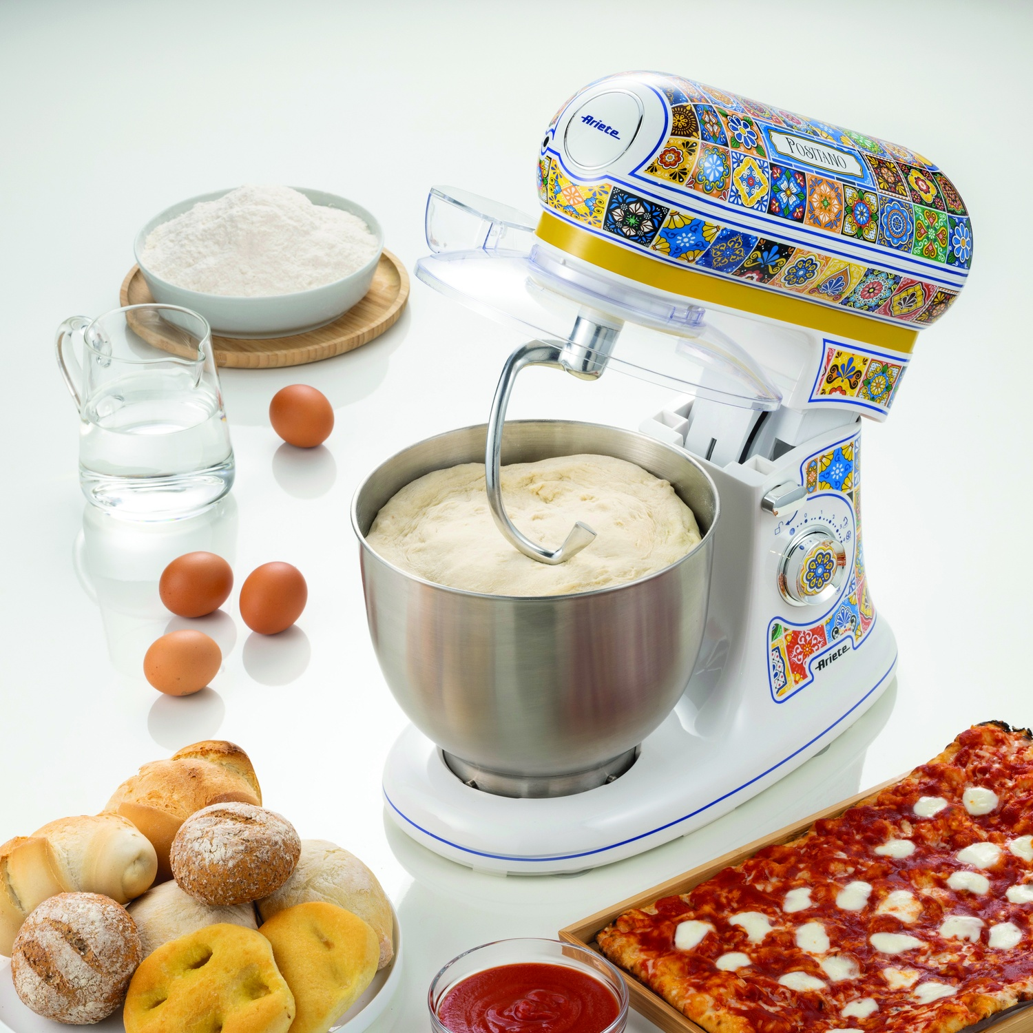 Un mixer da cucina con impasto dentro e ingredienti intorno: farina, acqua, uova, pane, pizza.