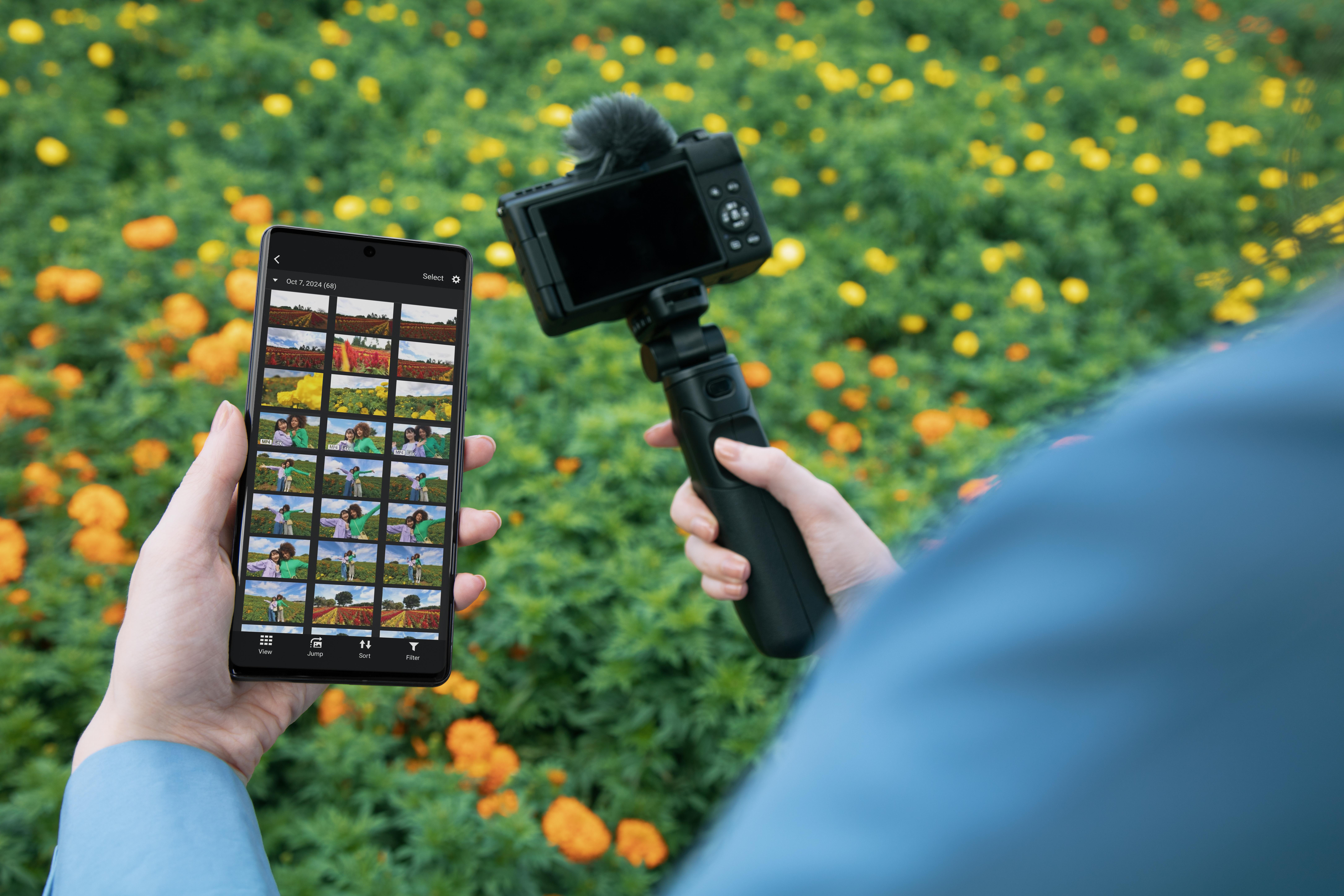 Eine Person hält ein Telefon mit Fotos und eine Kamera auf einem Stativ vor einem Blumenfeld.