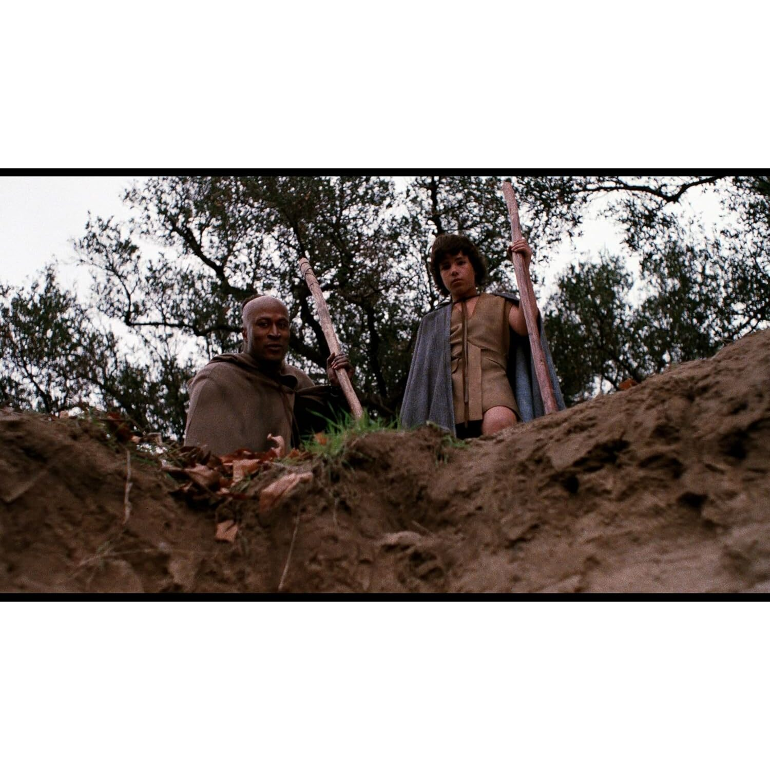 Two men in a field holding wooden poles. One is dark-skinned, the other is fair-skinned, in front of trees.
