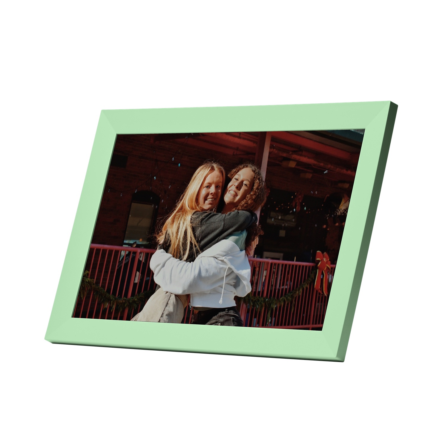 Two smiling women hugging in a green framed picture.  They are in front of a red building with a Christmas decoration.