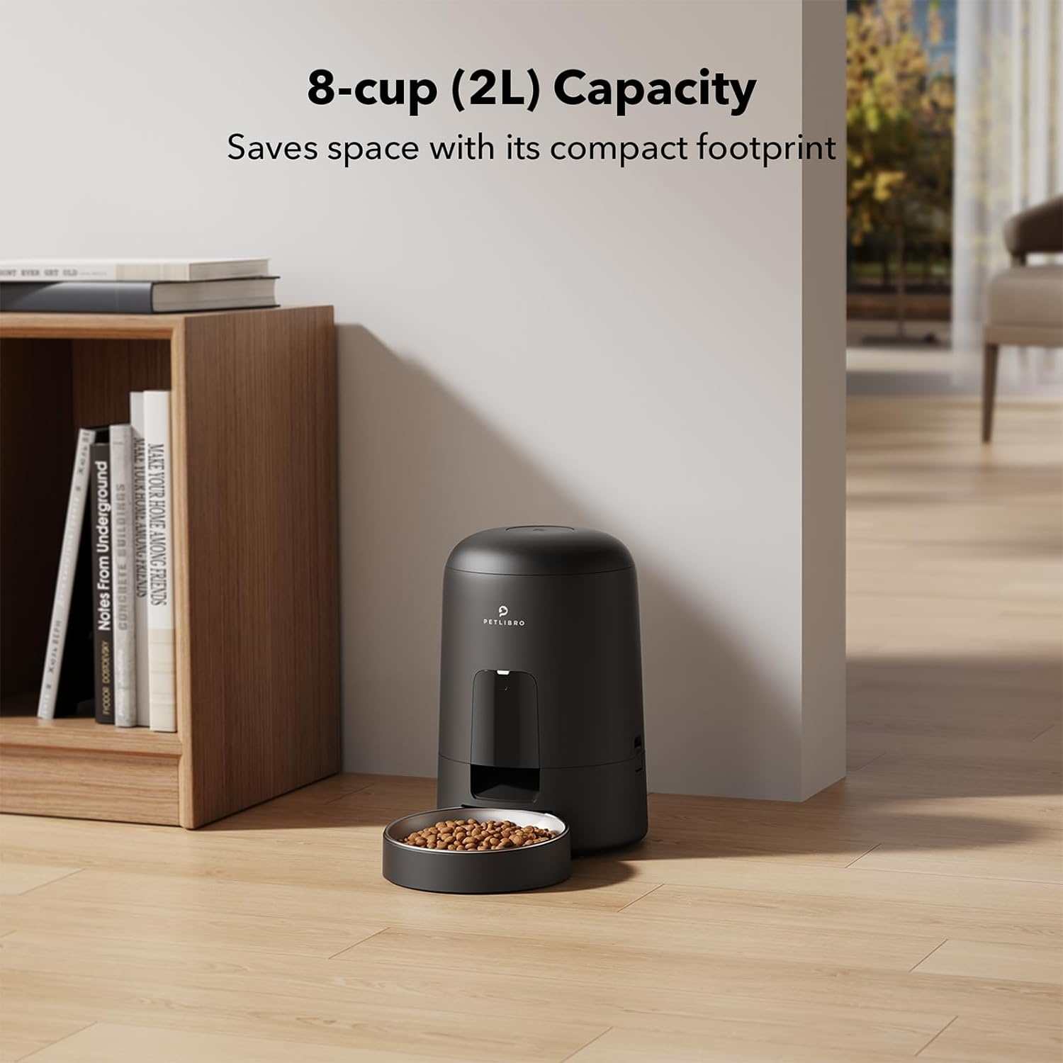 Black pet food dispenser with a bowl. A wooden shelf with books is in the background.
