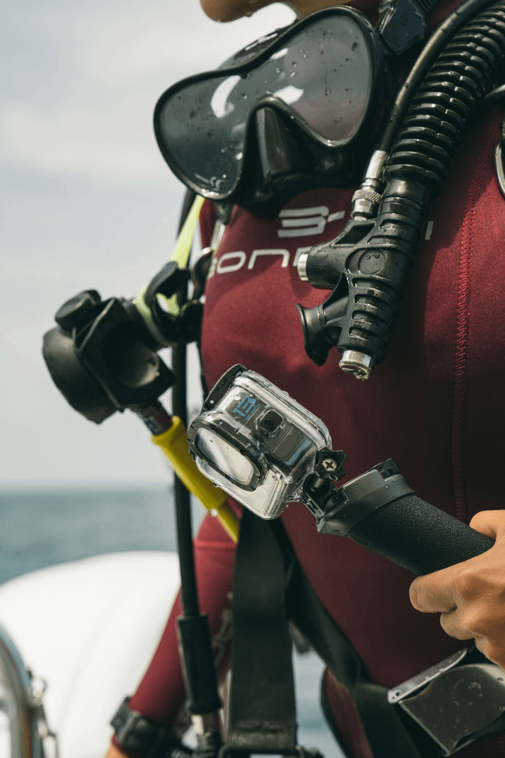 Un buzo con traje de neopreno granate sostiene una cámara. El equipo de buceo incluye máscara, regulador y una parte amarilla y negra. Se ven cielo y mar.