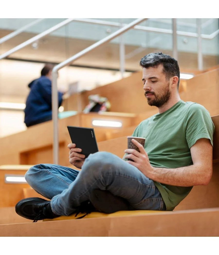 Un hombre sentado en escaleras, sosteniendo una tableta y una taza de café. Libros y otra persona al fondo.