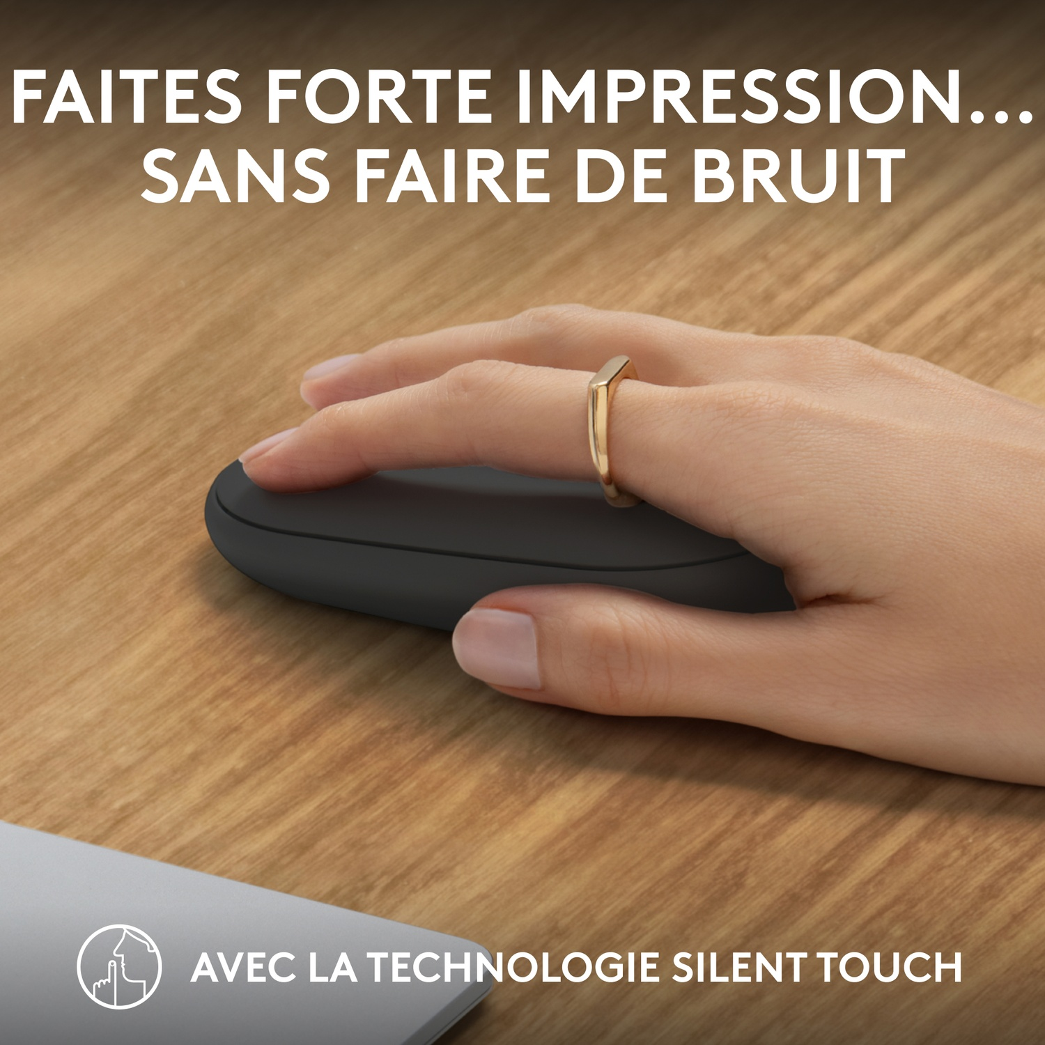 Une main portant une bague en or tient une souris noire ovale sur une surface en bois. Ordinateur portable gris en arrière-plan.