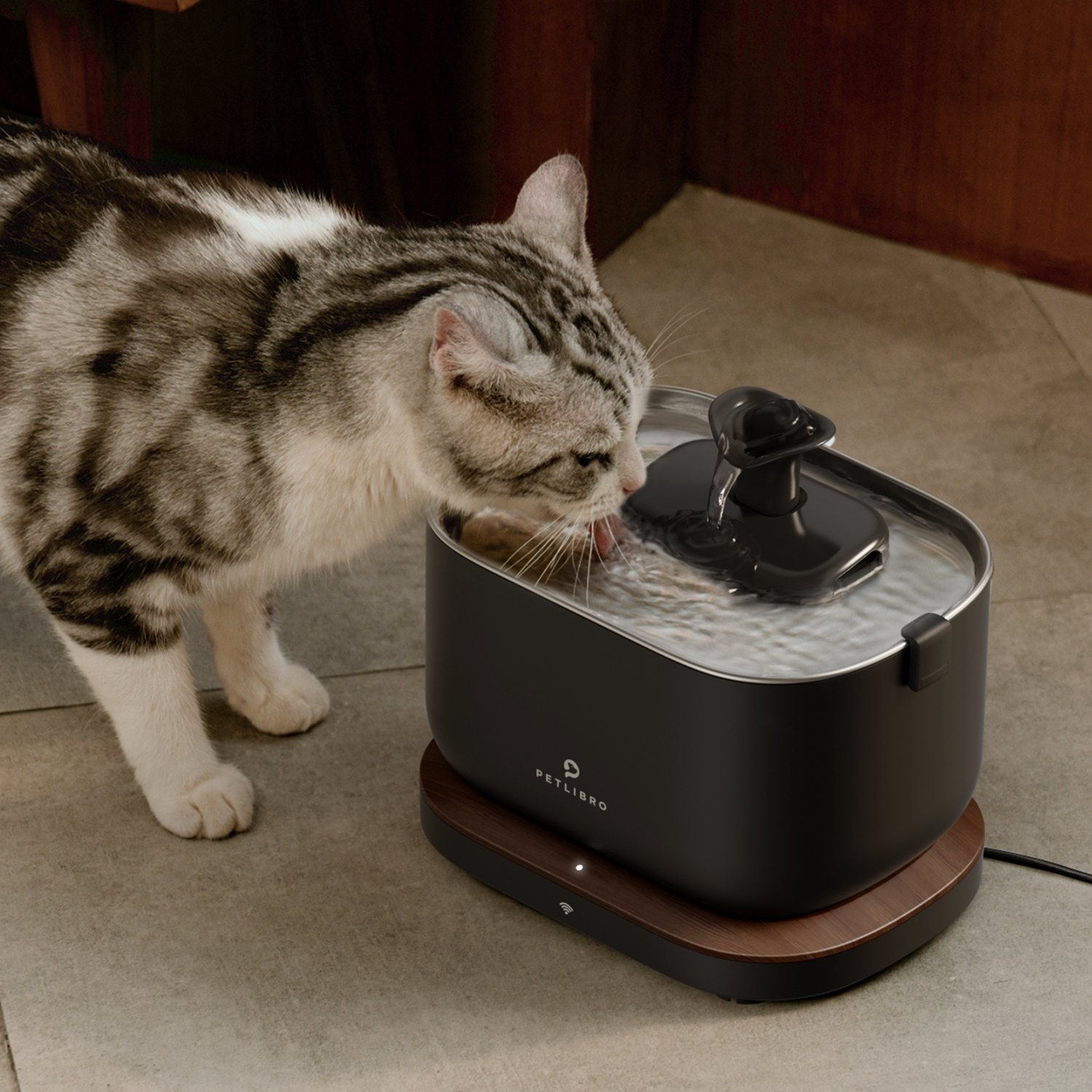 A cat drinks from a black pet water fountain. Wooden floor.