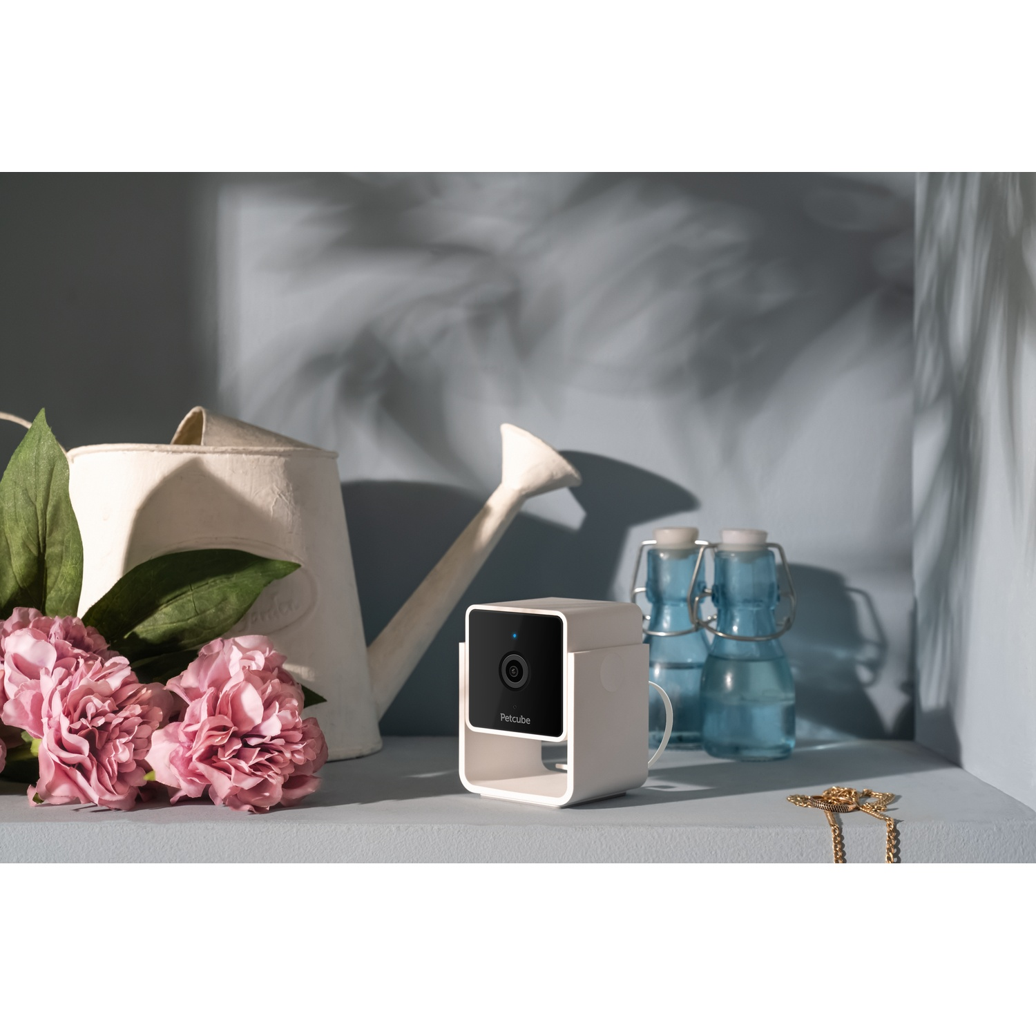 A Petcube camera on a shelf with flowers and bottles.