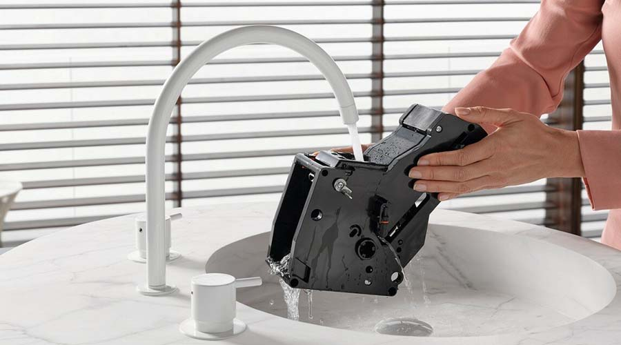 Osoba myje część ekspresu do kawy w zlewie z kranem. Białe tło.