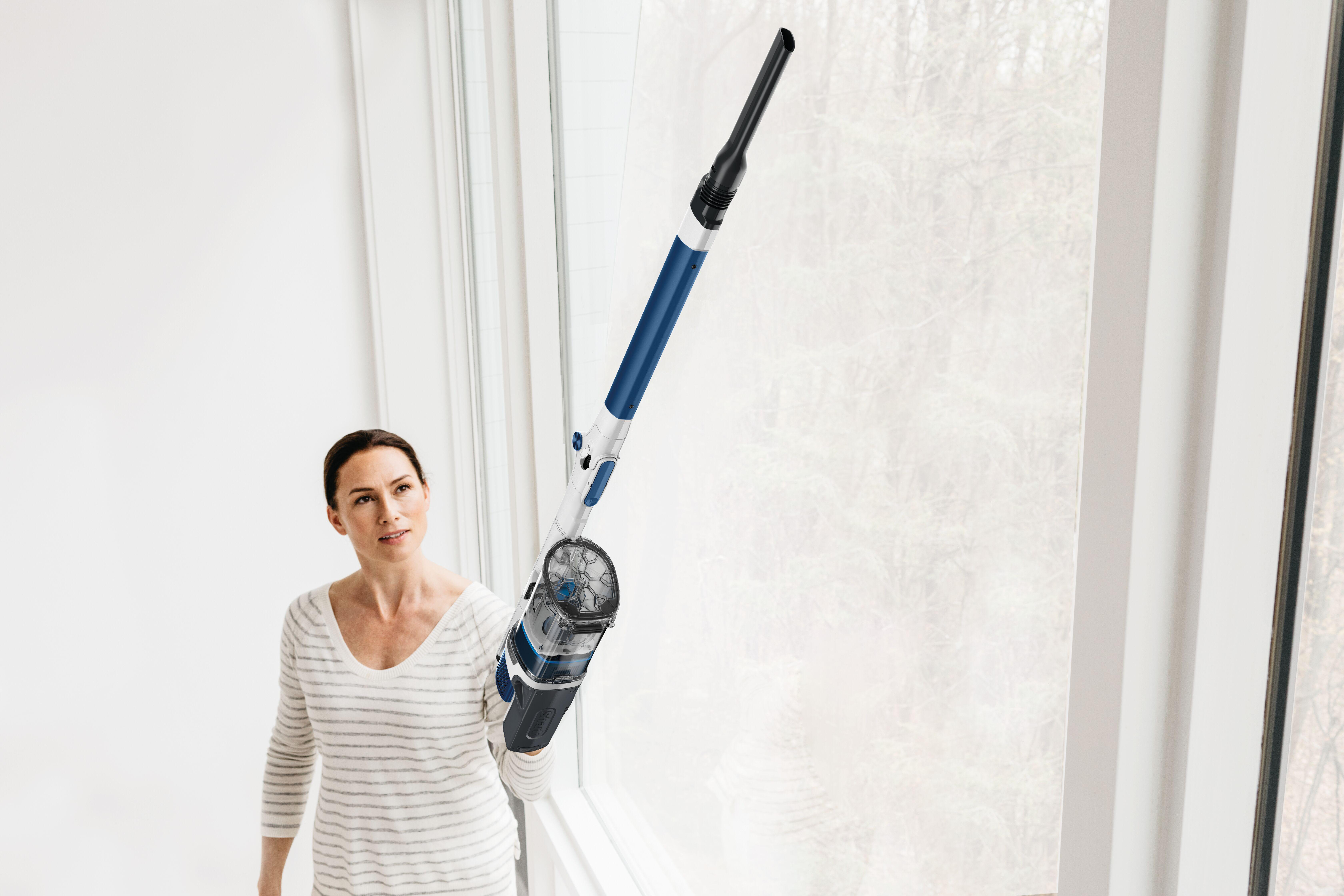 Frau reinigt ein Fenster mit einem kabellosen Staubsauger, heller und weißer Hintergrund.