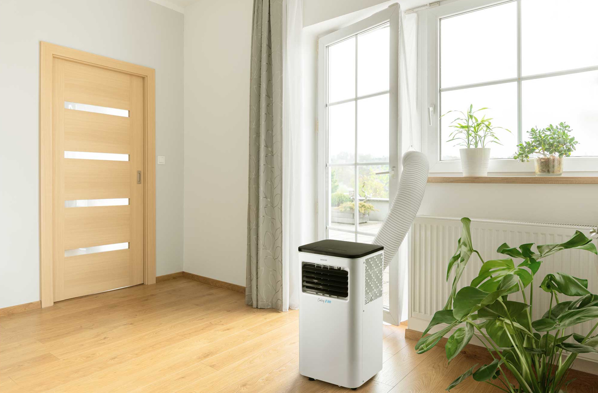 Biały klimatyzator przy oknie. Otwarte drzwi i rośliny. Drewniana podłoga.