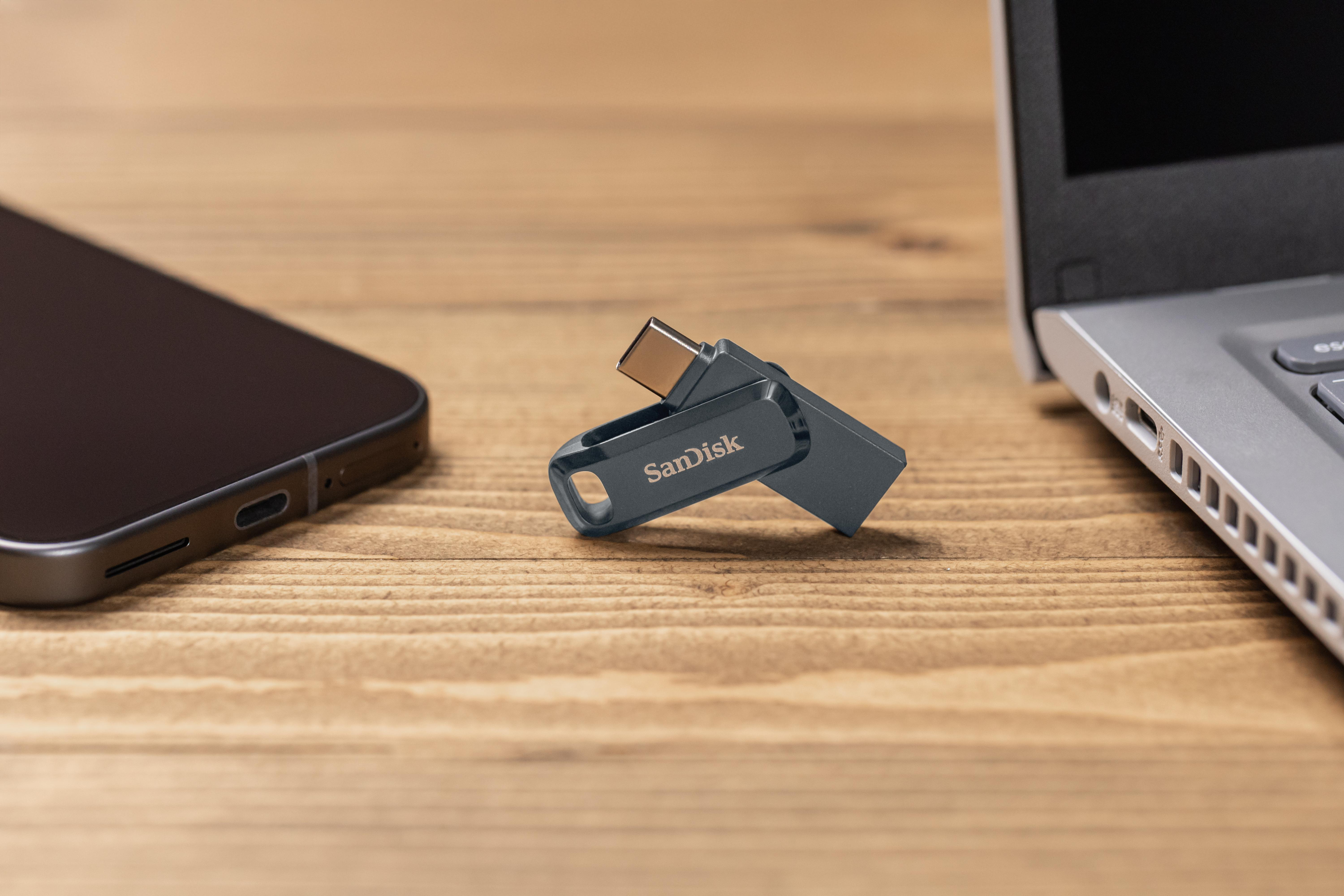 USB-Stick zwischen Telefon und Laptop auf einem Holztisch. Sandisk-Logo sichtbar.