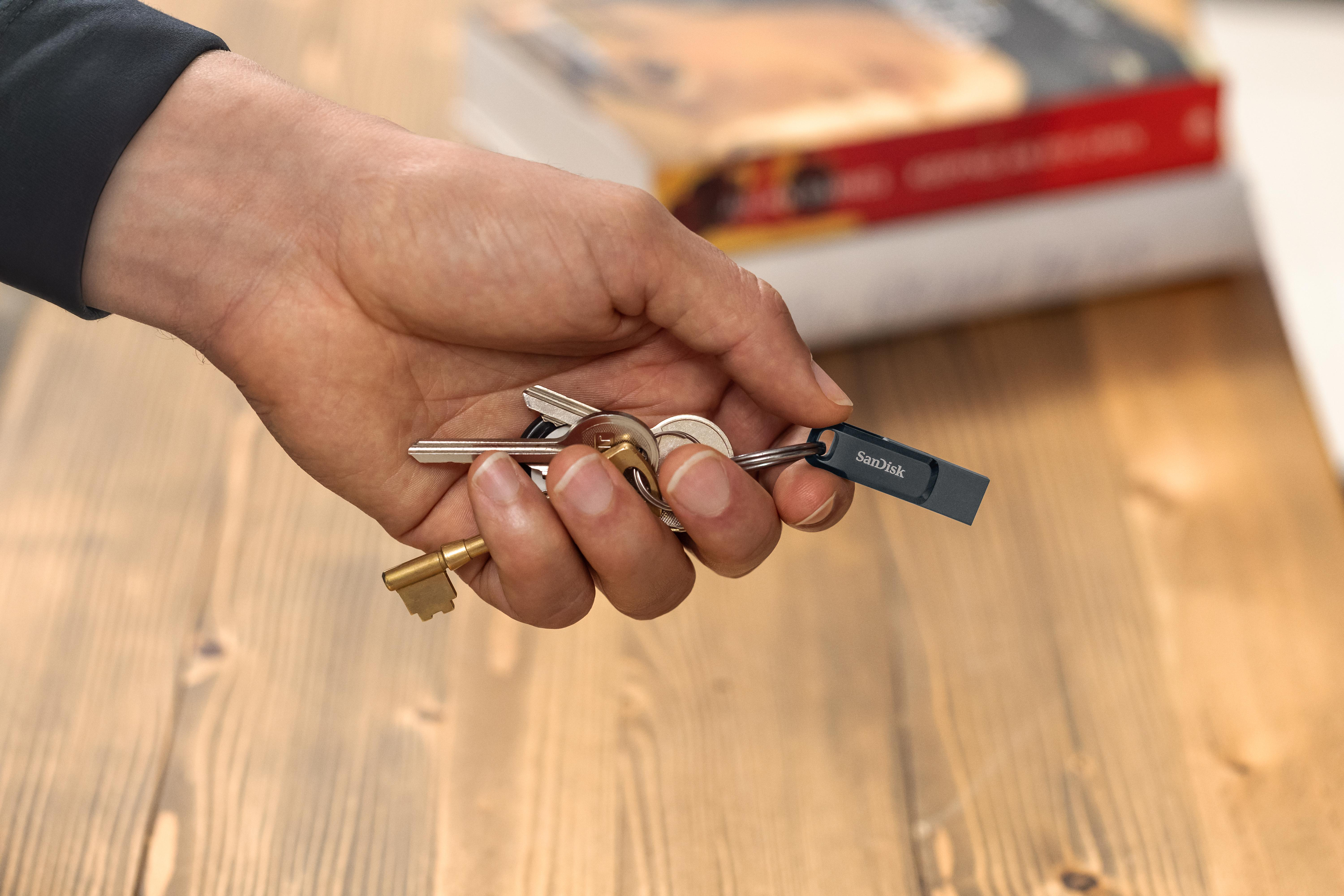 Hand hält Schlüssel und einen Sandisk USB-Stick auf Holztisch. Bücher im Hintergrund.