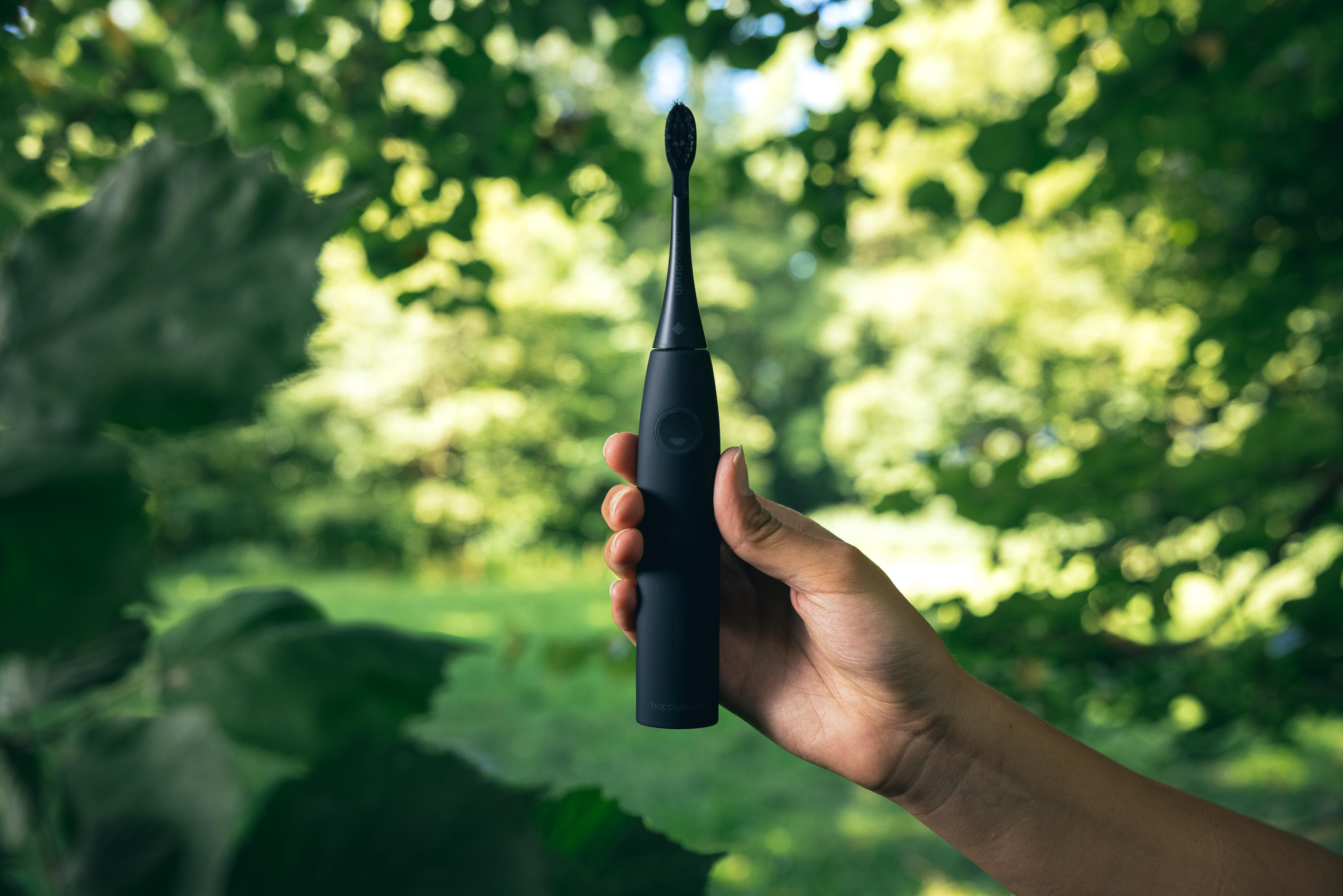 Eine Hand, die eine schwarze elektrische Zahnbürste vor einem unscharfen grünen Hintergrund hält.