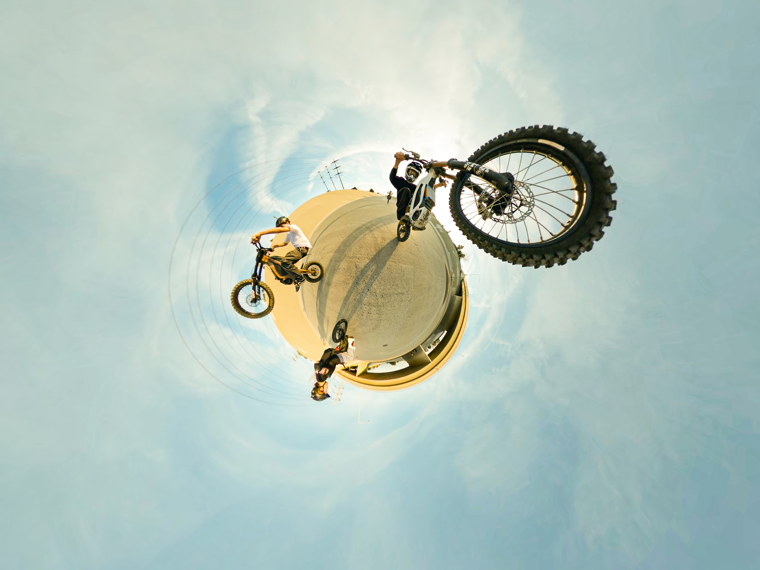 Motocicletas haciendo acrobacias en el aire. Un piloto está boca abajo. El cielo es azul con nubes.