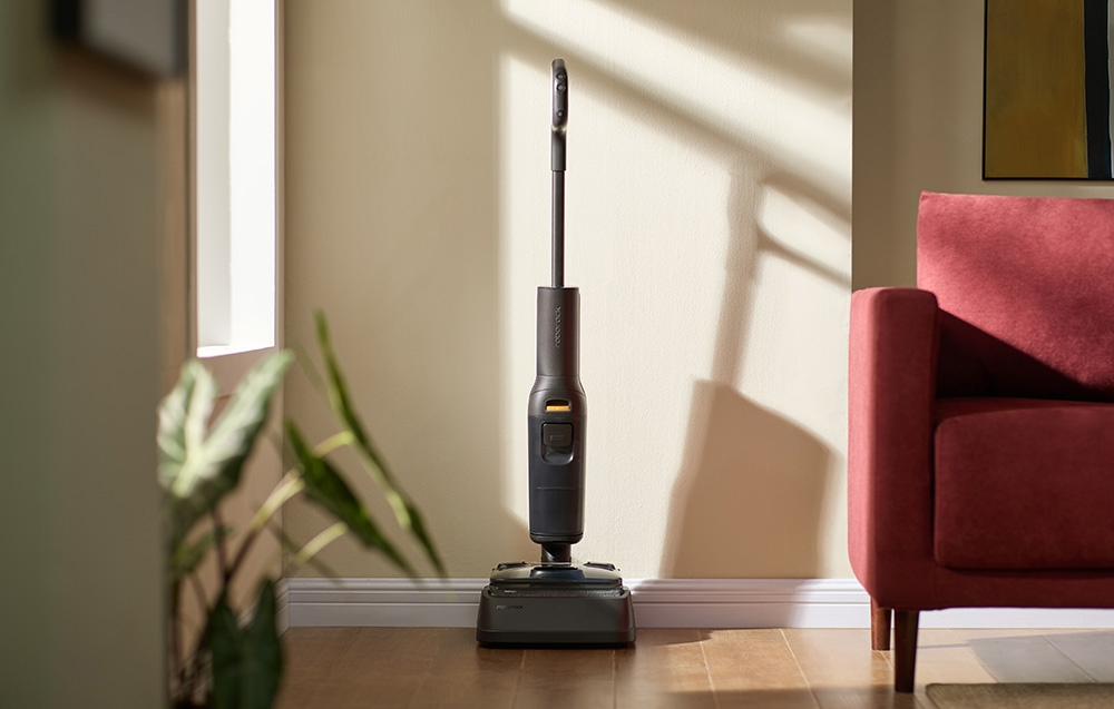 Czarno-szary odkurzacz bezprzewodowy stoi na jasnej drewnianej podłodze. Czerwona kanapa w tle.
