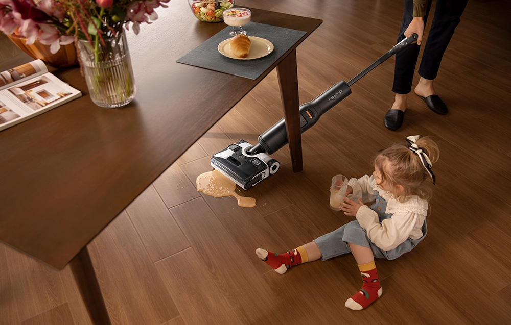 Kobieta odkurza rozlaną ciecz obok dziecka na podłodze. Stół i jedzenie w tle.