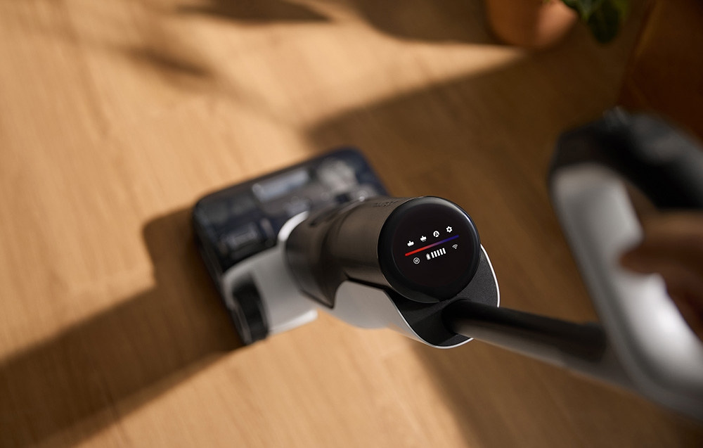 Głowica odkurzacza na drewnie. Wyświetlacz pokazuje baterię i ustawienia. Ręka w pobliżu. Tło rozmyte.