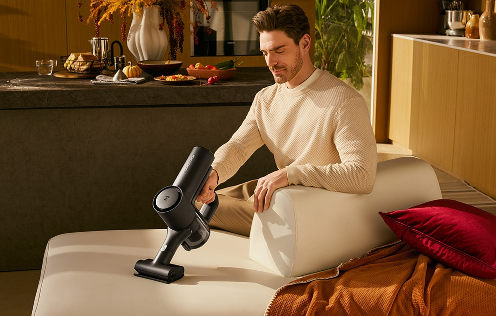 Mężczyzna odkurza kanapę odkurzaczem ręcznym. W tle ciemna wyspa kuchenna.