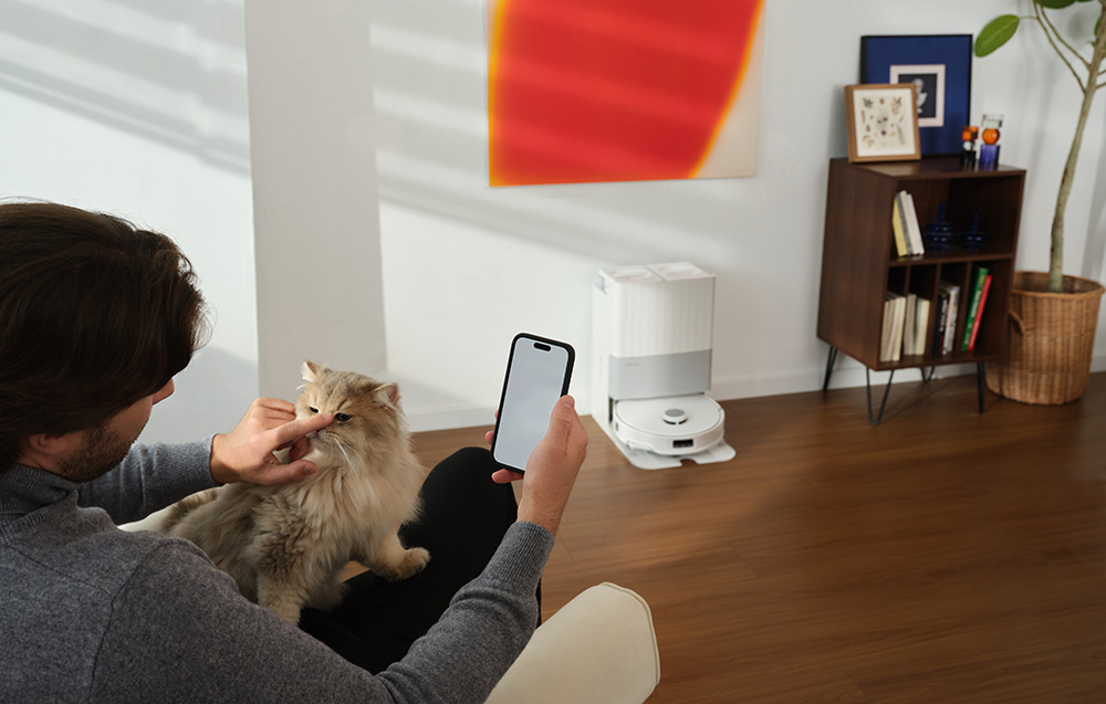 Mężczyzna z kotem na kolanach wchodzi w interakcję z telefonem, w tle robot sprzątający.
