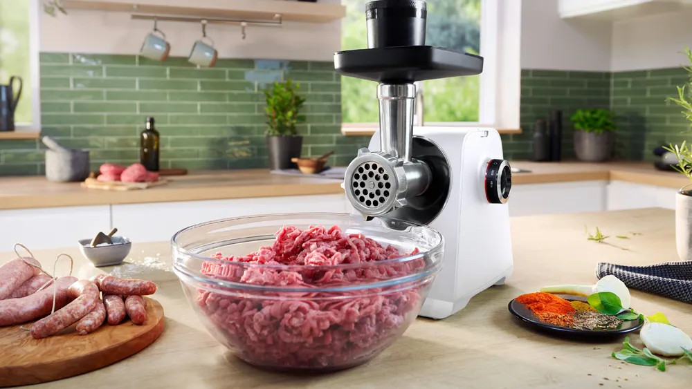 Maszynka do mięsa z mielonym mięsem w szklanej misce, otoczona składnikami.