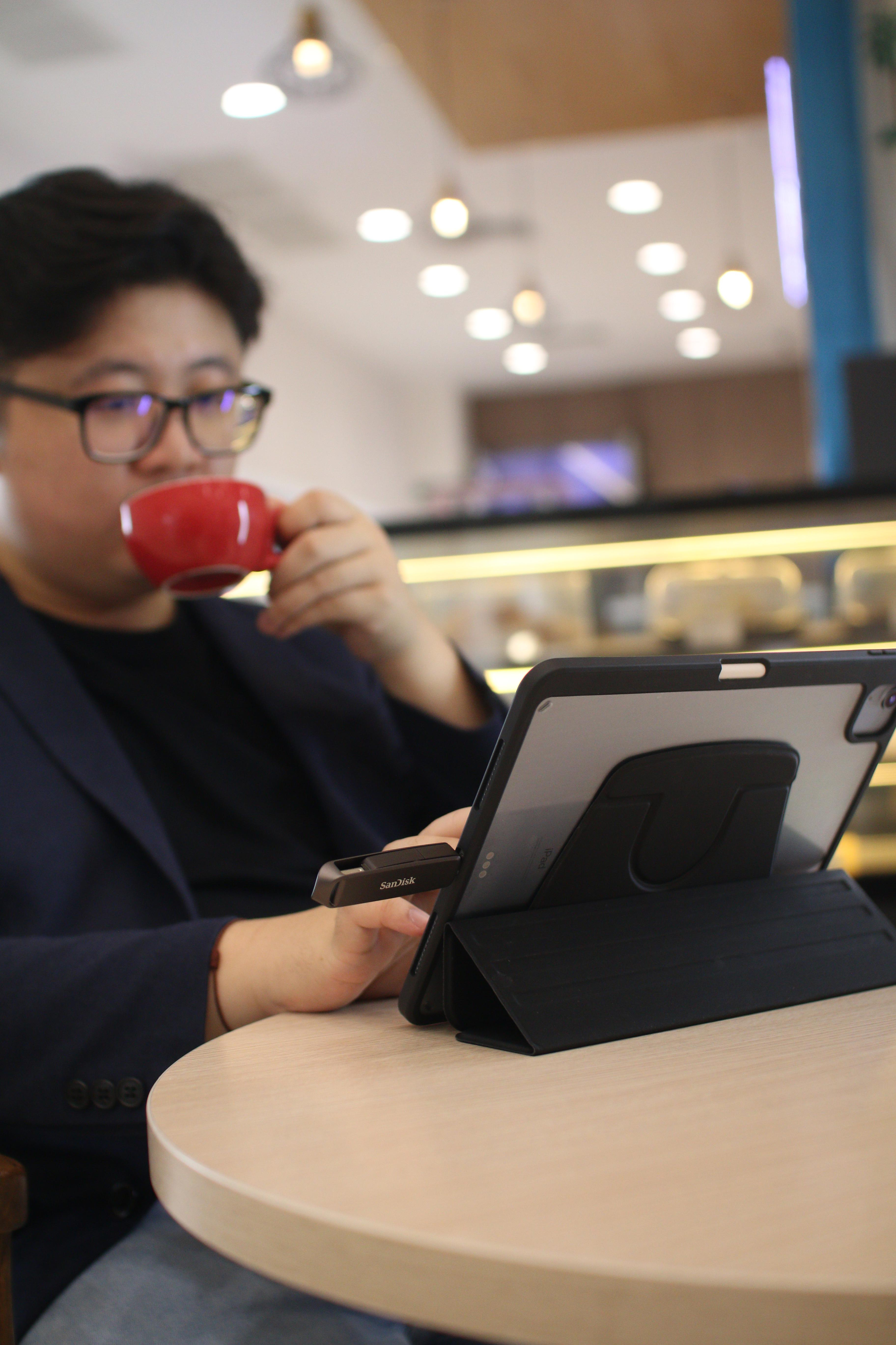 Ein Mann hält ein Tablet, trinkt aus einer roten Tasse an einem Tisch.