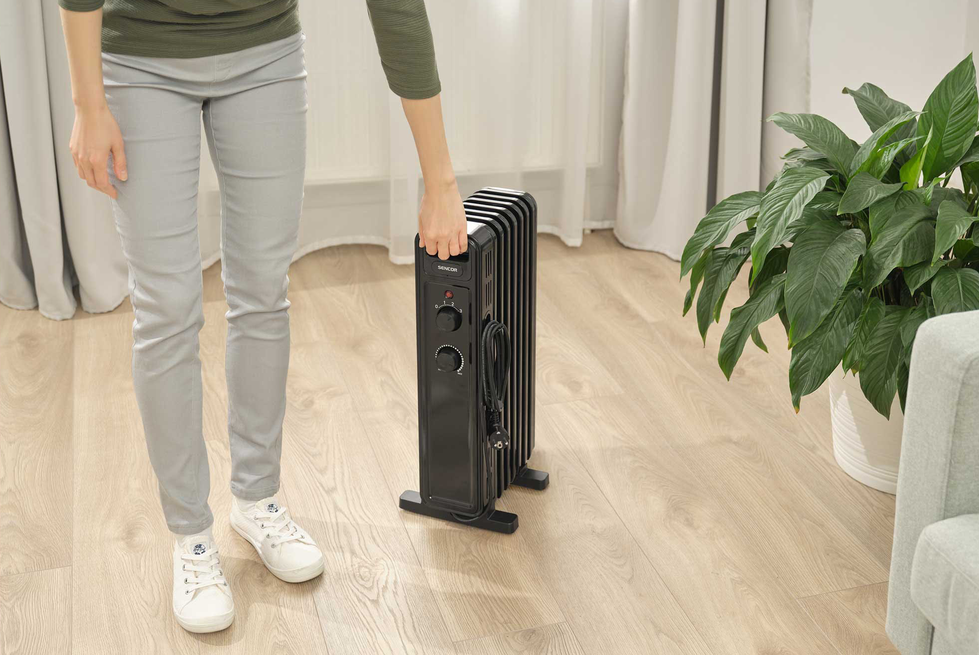 Egy személy fekete olajradiátort tart. Egy fa padlón állnak egy növény közelében.