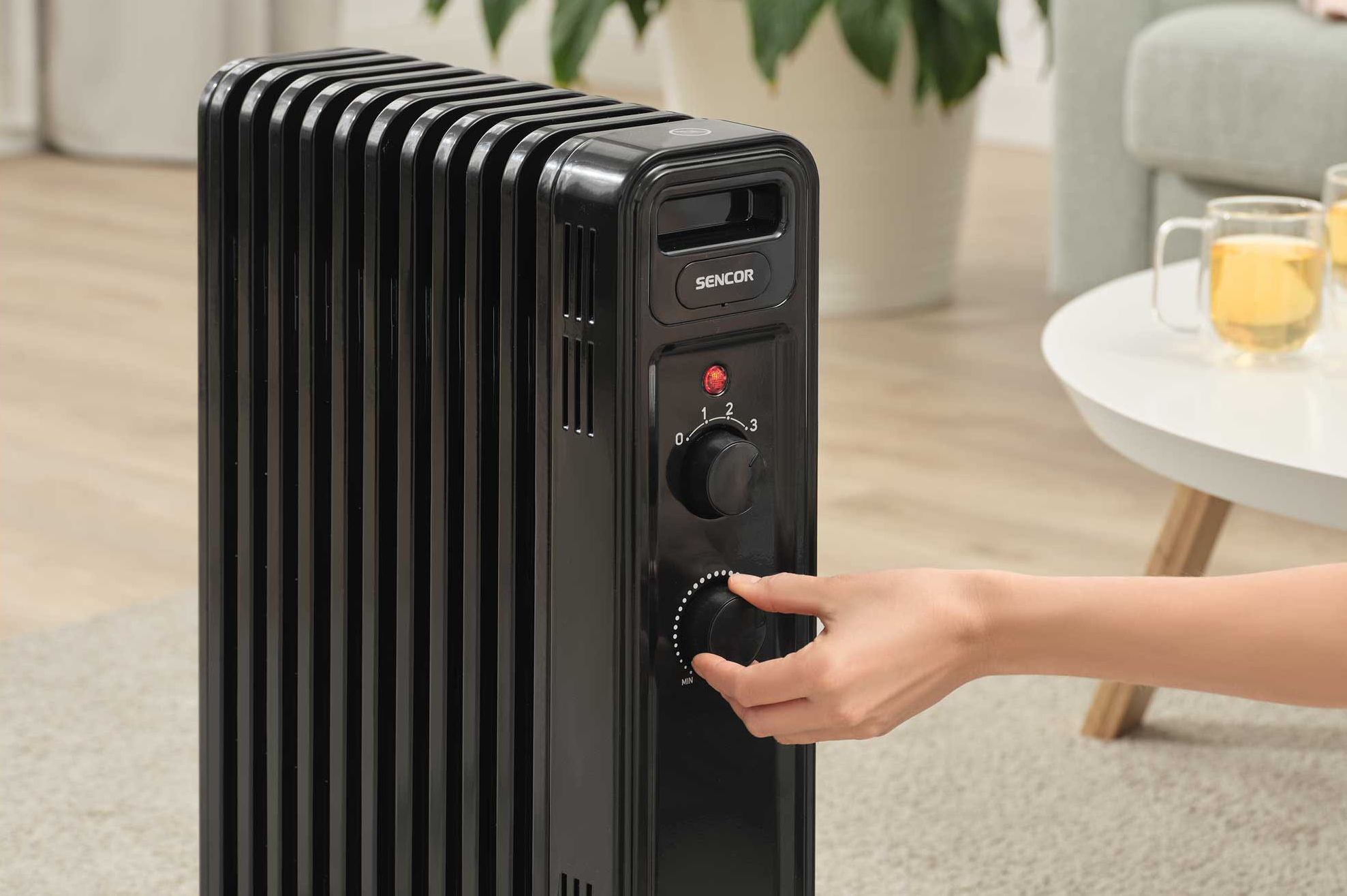 Egy kéz egy fekete elektromos olajradiátort állít be. Egy fehér asztal, egy kanapé és egy háttér.