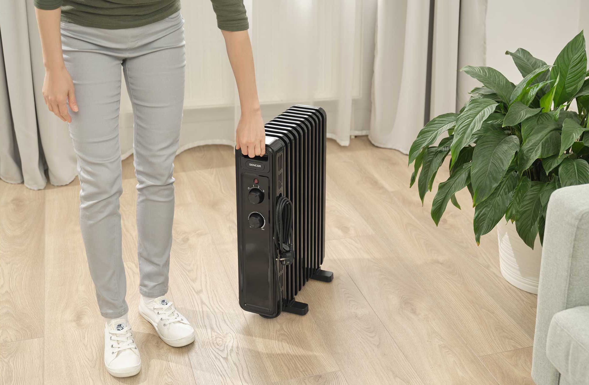 Egy személy fekete olajradiátort tart, fa padlón áll, egy fehér cseréppel és egy kanapéval.