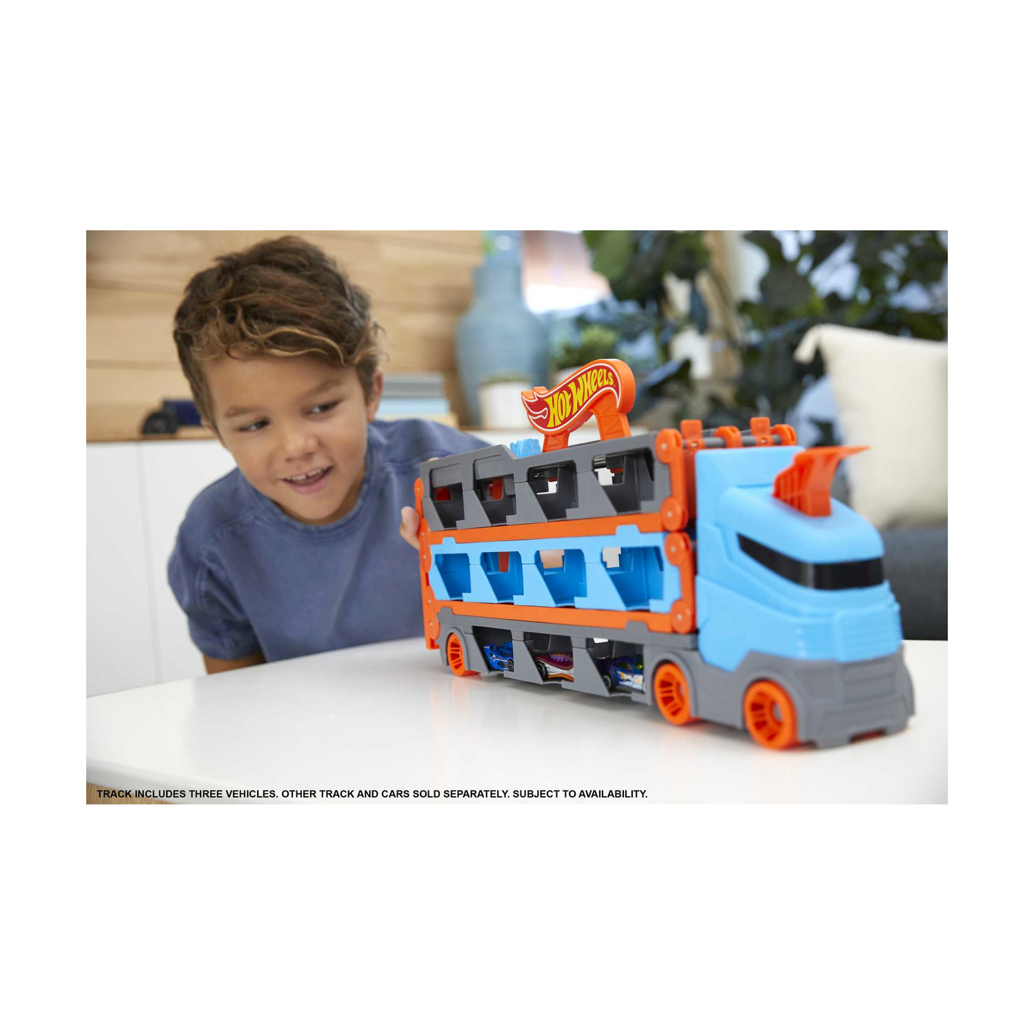Un bambino guarda un camion Hot Wheels blu e arancione. Il camion contiene macchinine. Lo sfondo presenta un tavolo e una finestra.