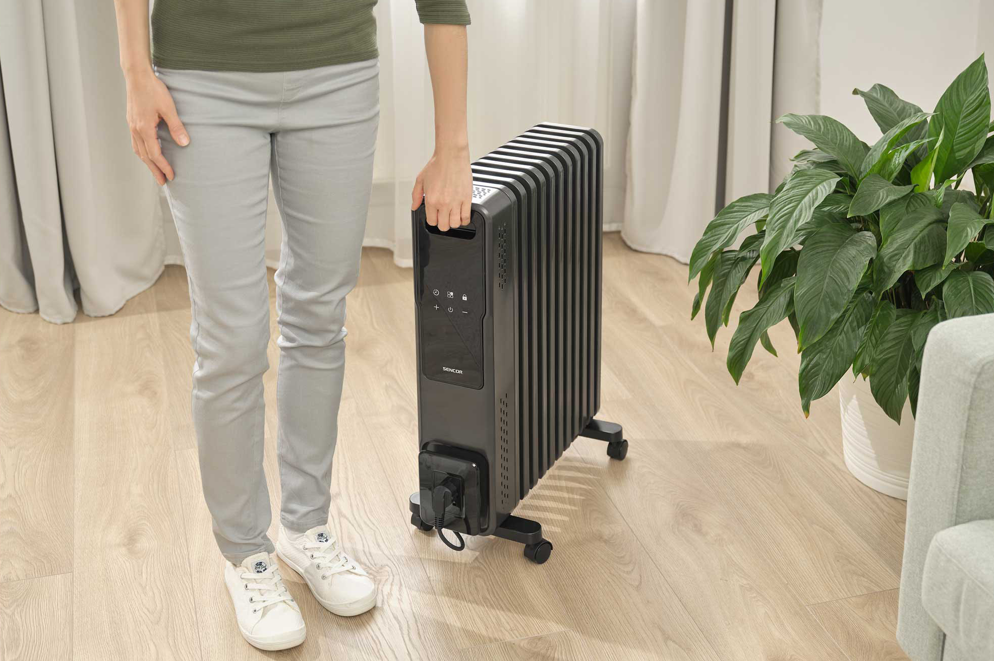 Egy nő fekete elektromos fűtőtestet tart. Háttérben szobanövény és kanapé.