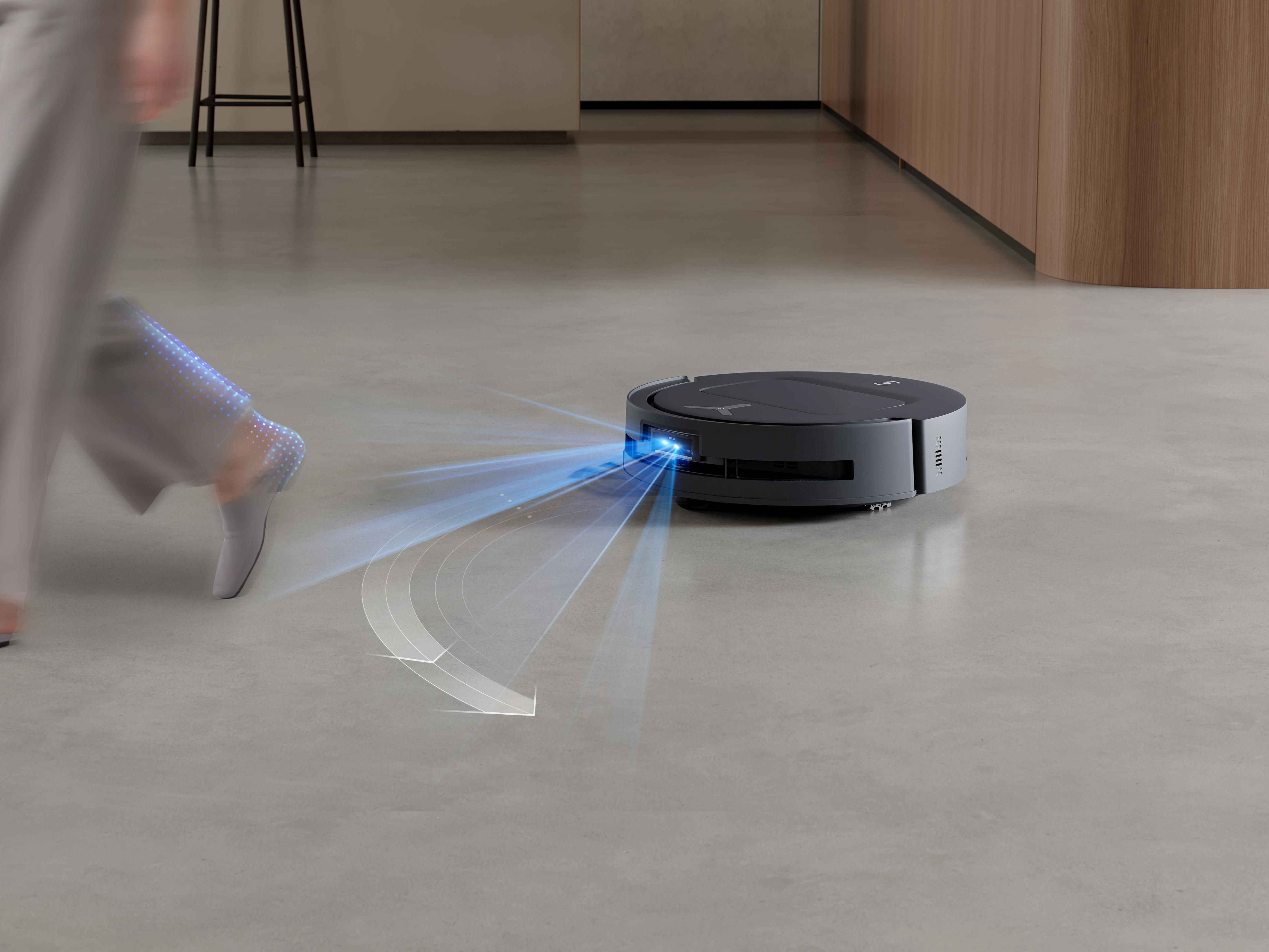 Ein Roboter-Staubsauger reinigt einen Boden. Beine einer Person im Hintergrund.