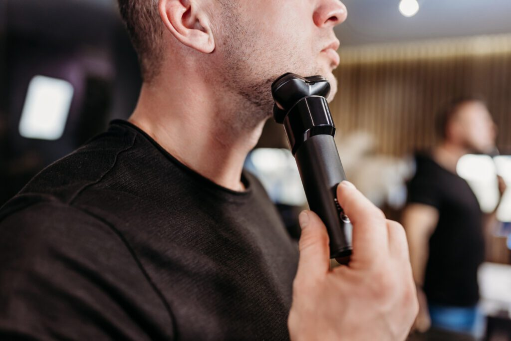 Mężczyzna goli brodę golarką elektryczną. Tło jest rozmyte.