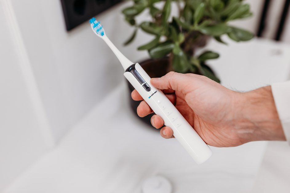 Biała elektryczna szczoteczka do zębów w dłoni. Łazienka z rośliną w tle.