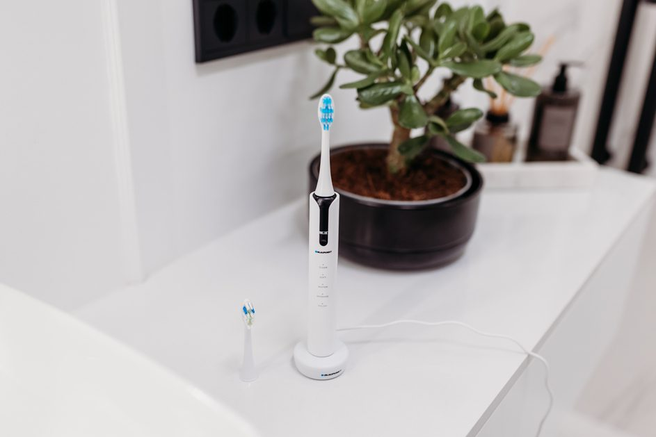 Elektryczna szczoteczka do zębów na białym blacie. Mała roślina w tle. Szczoteczka jest podłączona.