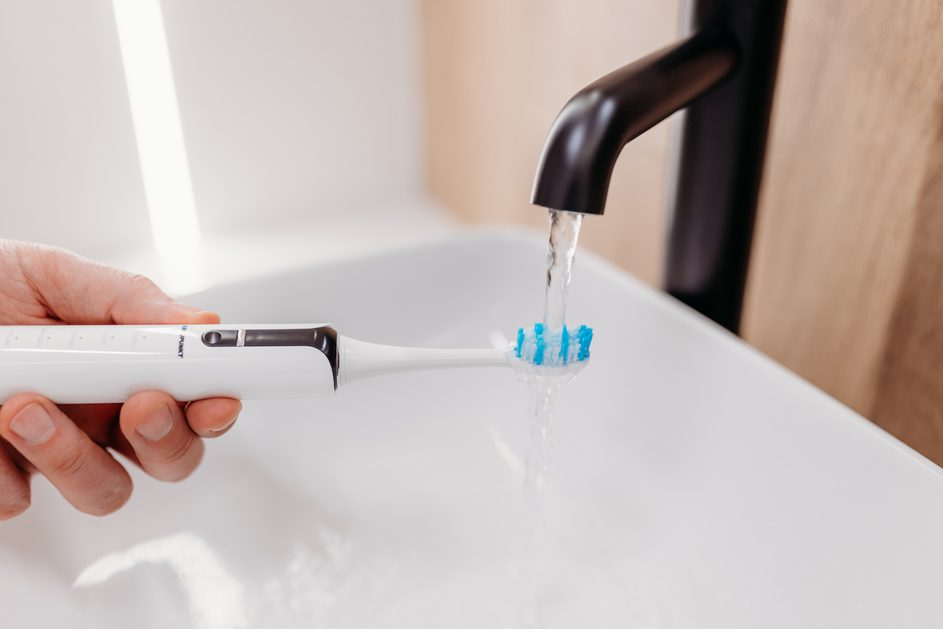 Ręka trzyma białą elektryczną szczoteczkę do zębów pod kranem. Woda spływa po niebieskim włosiu.