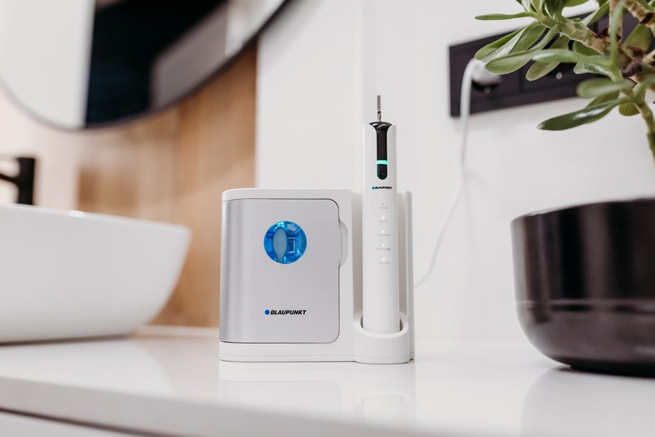 Biała szczoteczka do zębów i stacja ładująca na białej powierzchni. Lustro i roślina w tle.