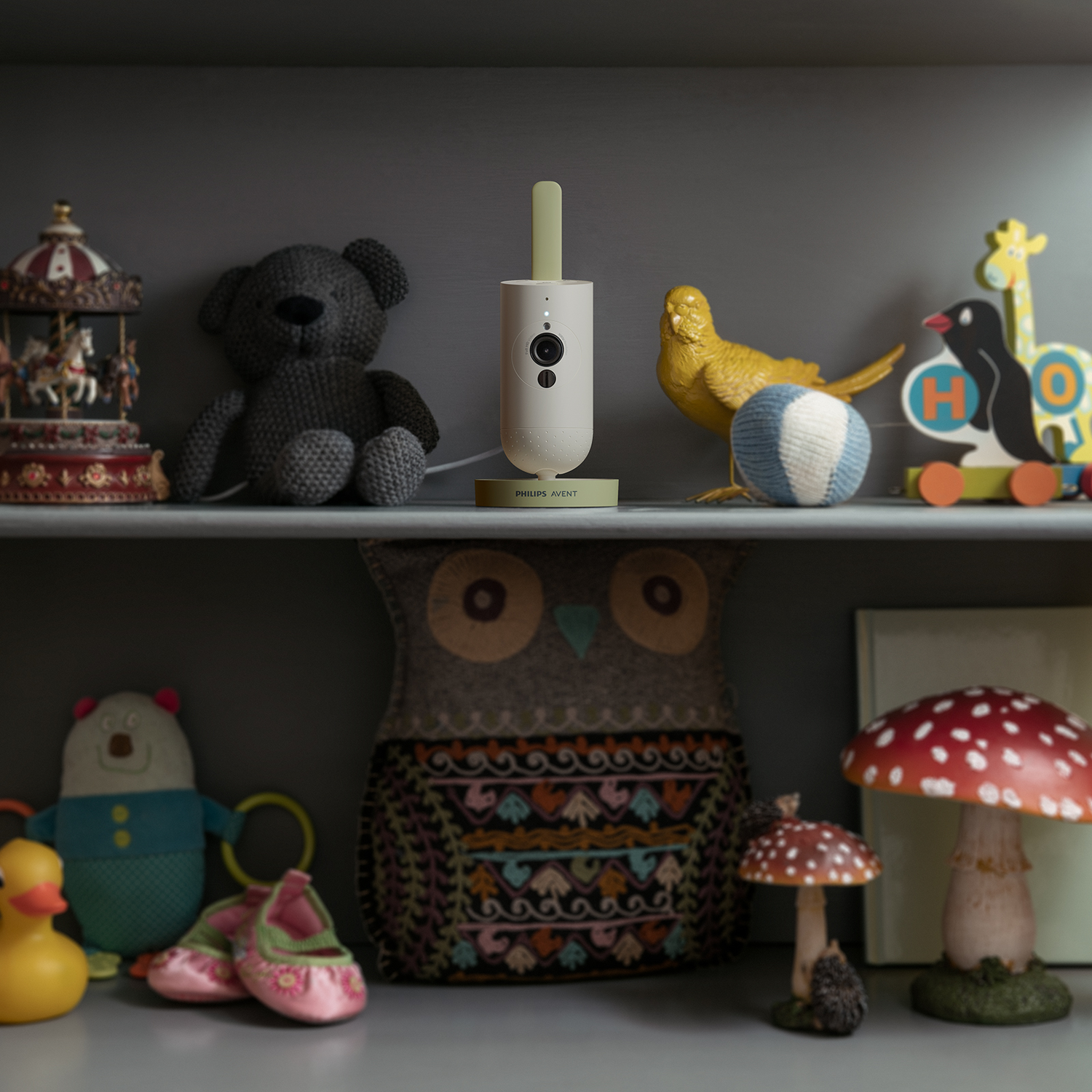 Een babyfoon op een plank met speelgoed, waaronder een teddybeer, vogel en uil decoratie.