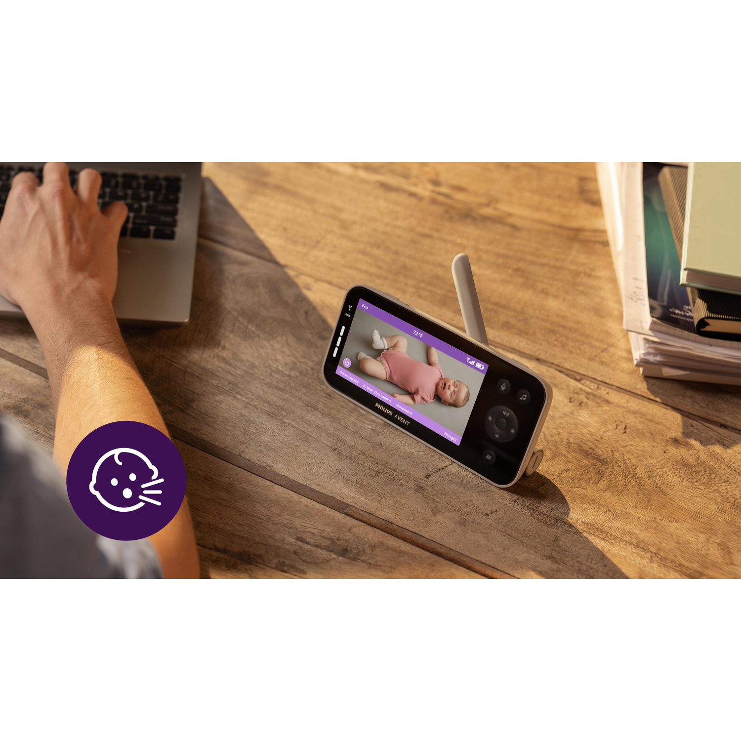 Een scherm van een babyfoon, met een baby. Een hand houdt de monitor vast. Laptop en papieren op de achtergrond.