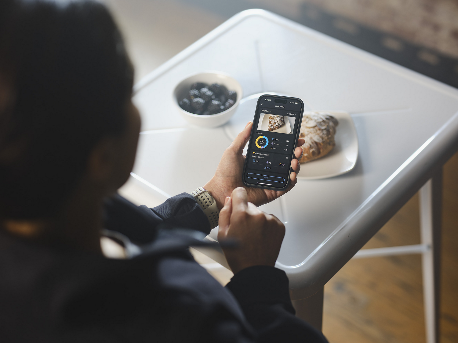 Persona mirando un smartphone mostrando detalles de comida. Un bol de moras y un pastel presentes.