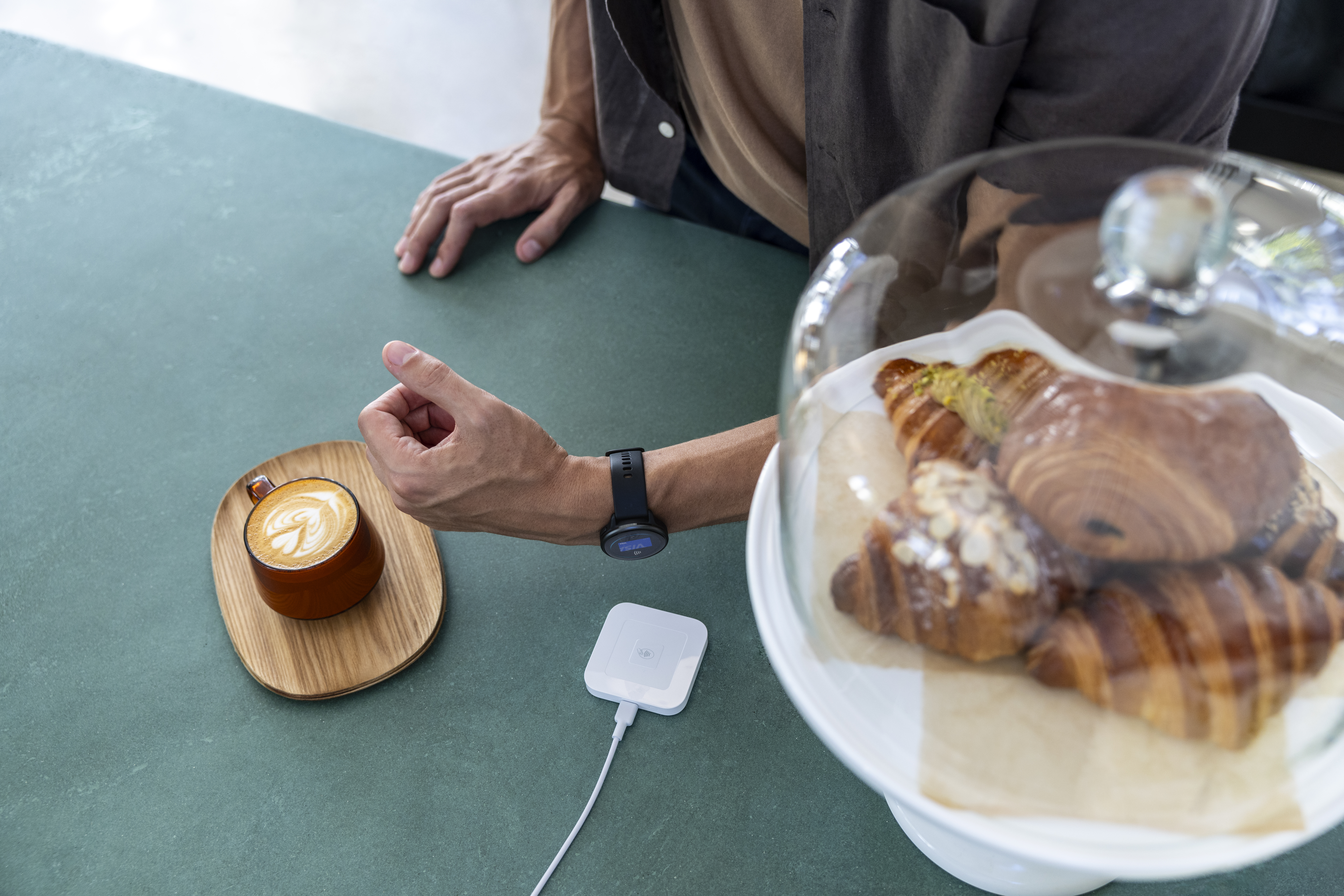 Person mit Smartwatch und Kaffee auf dem Tisch, Gebäck unter Glasabdeckung.