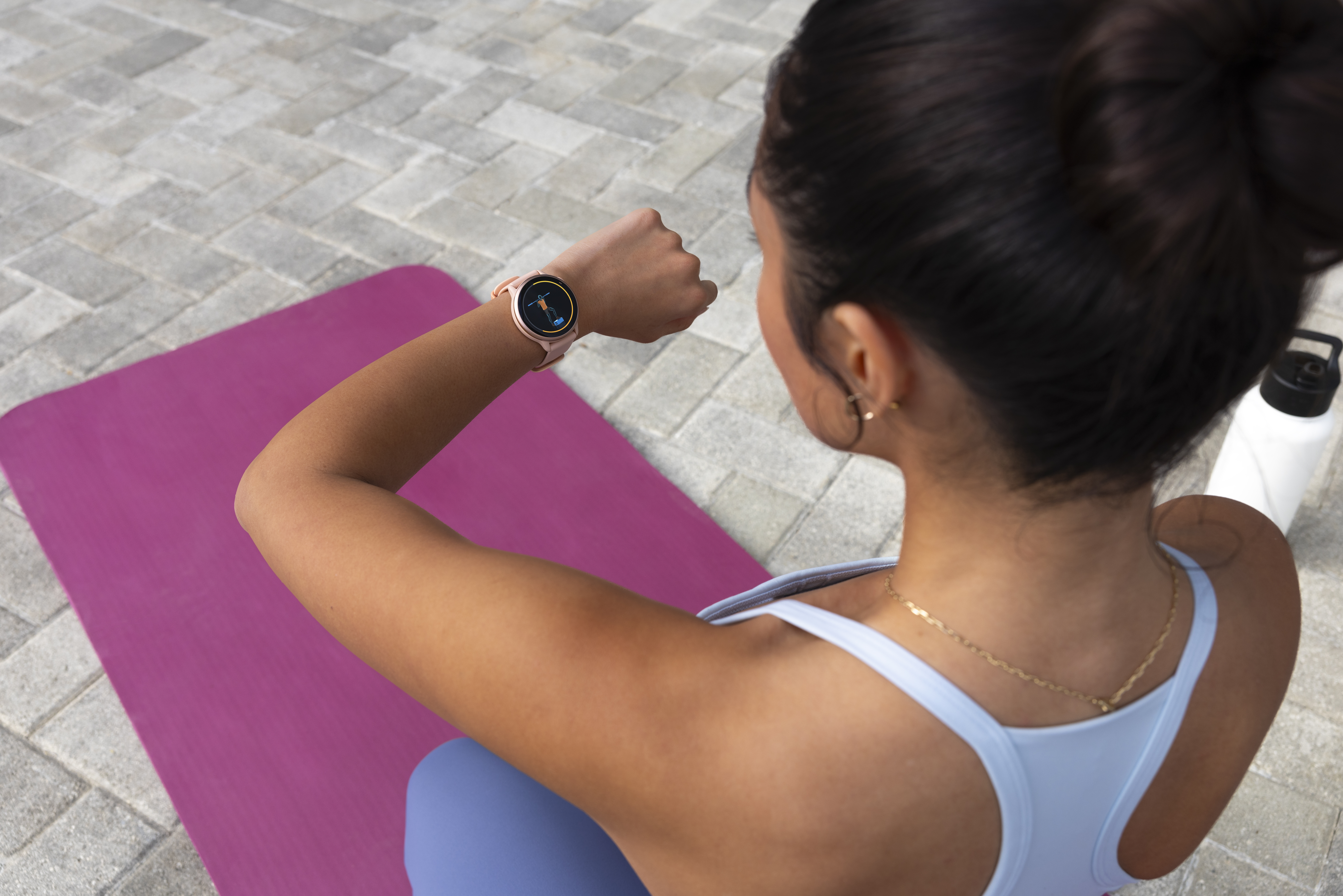 Frau, die ihre Smartwatch auf einer rosa Yogamatte überprüft, mit einer Wasserflasche und einem hellblauen Top.